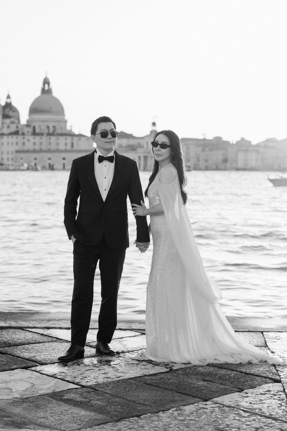 Elegant couple in wedding attire holding hands on Venice waterfront with Santa Maria della Salute in black and white.