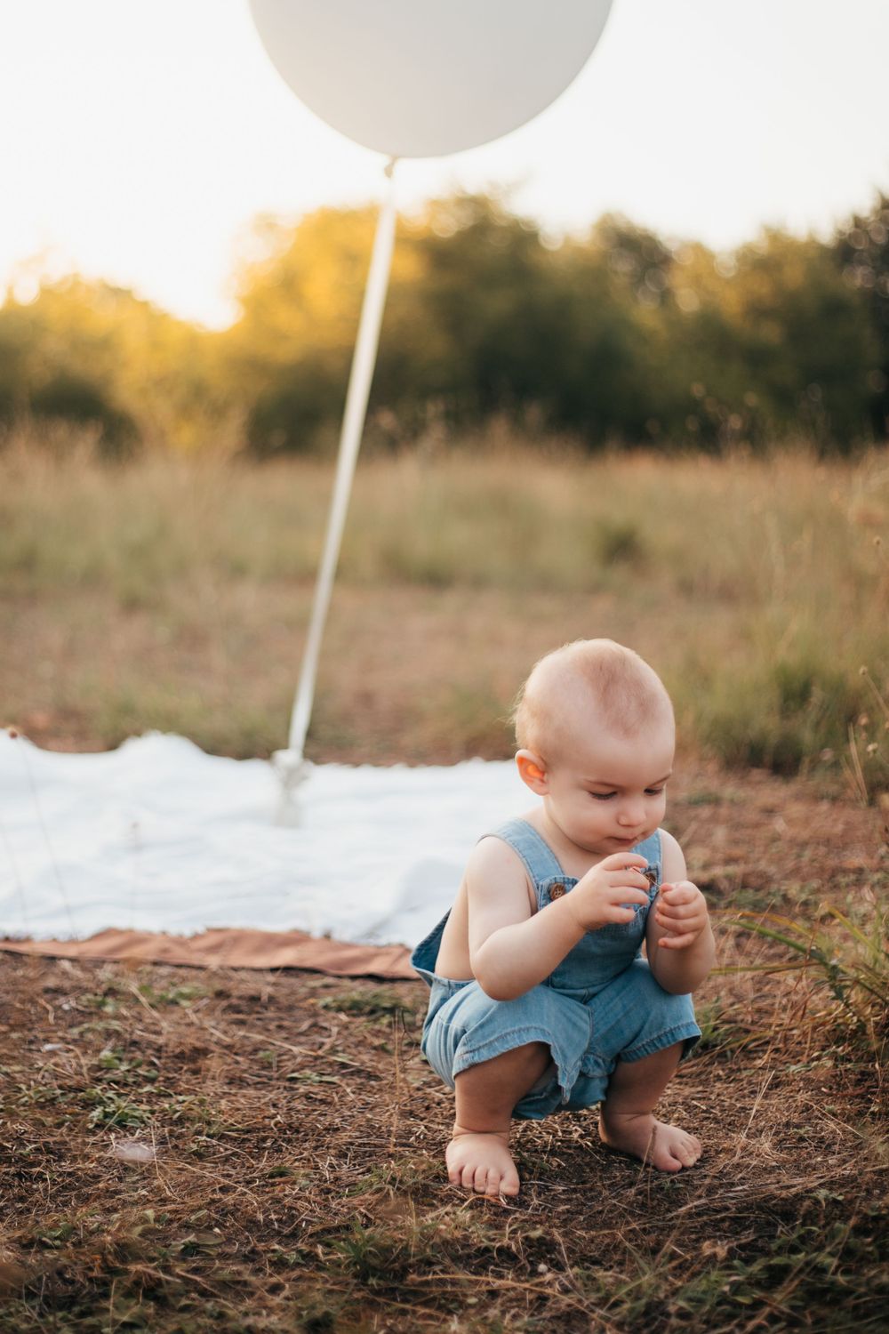 Small child in blue overalls crouches near large white balloon on white blanket during sunset.