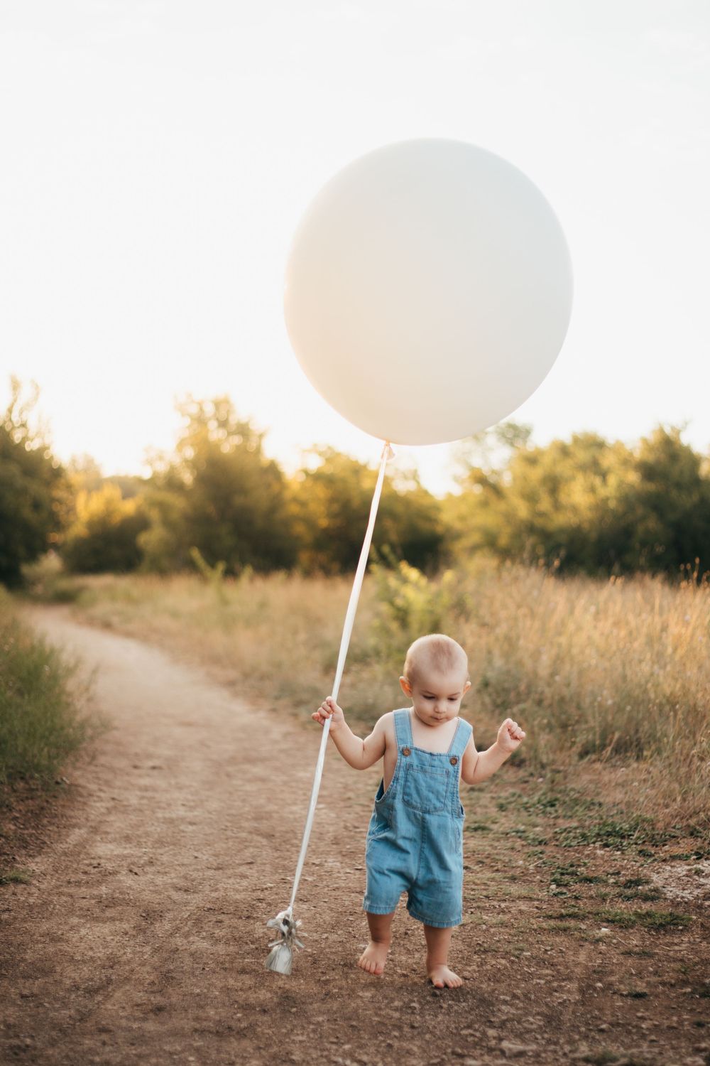 Toddler in denim overalls walks barefoot on dirt path holding large white balloon.