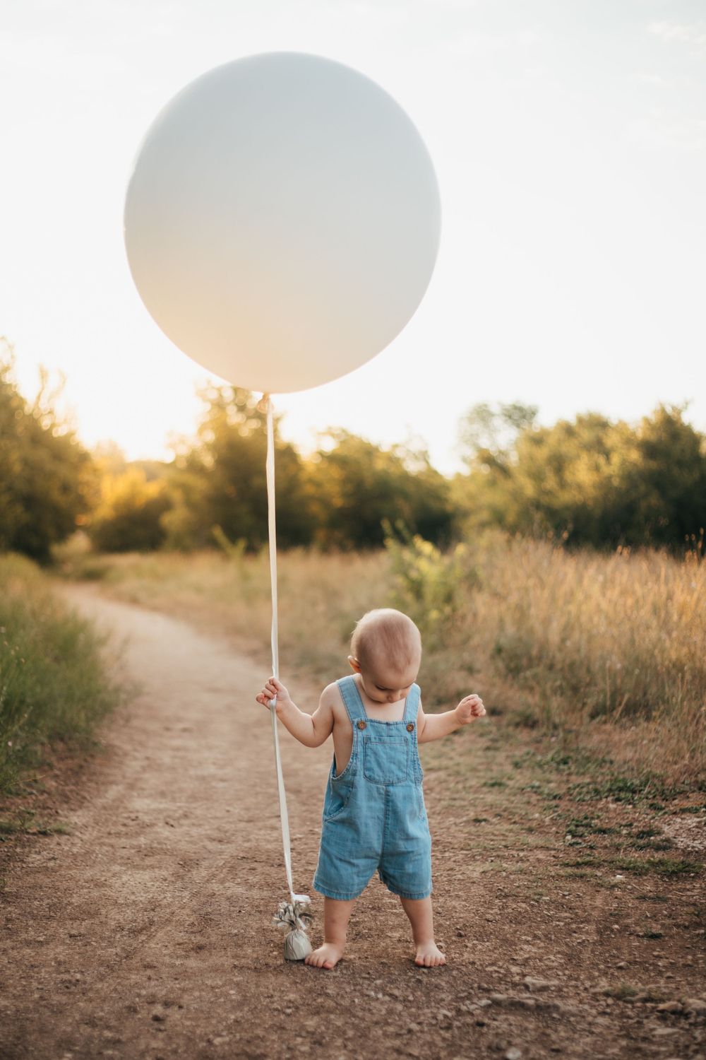Small child in blue overalls stands on dirt path holding giant white balloon at sunset.