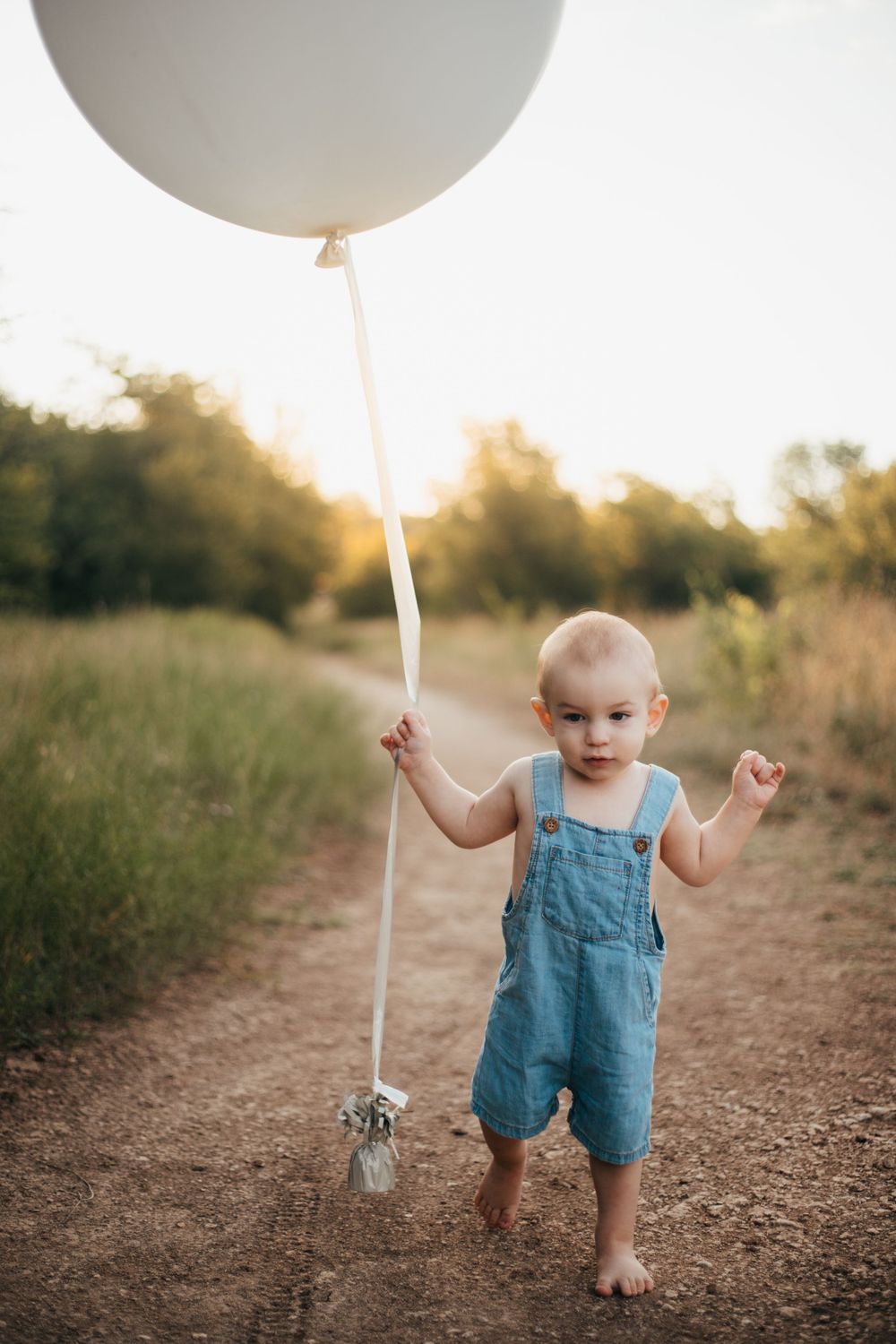 A toddler in blue overalls walks barefoot on a dirt path while holding a large white balloon at sunset.
