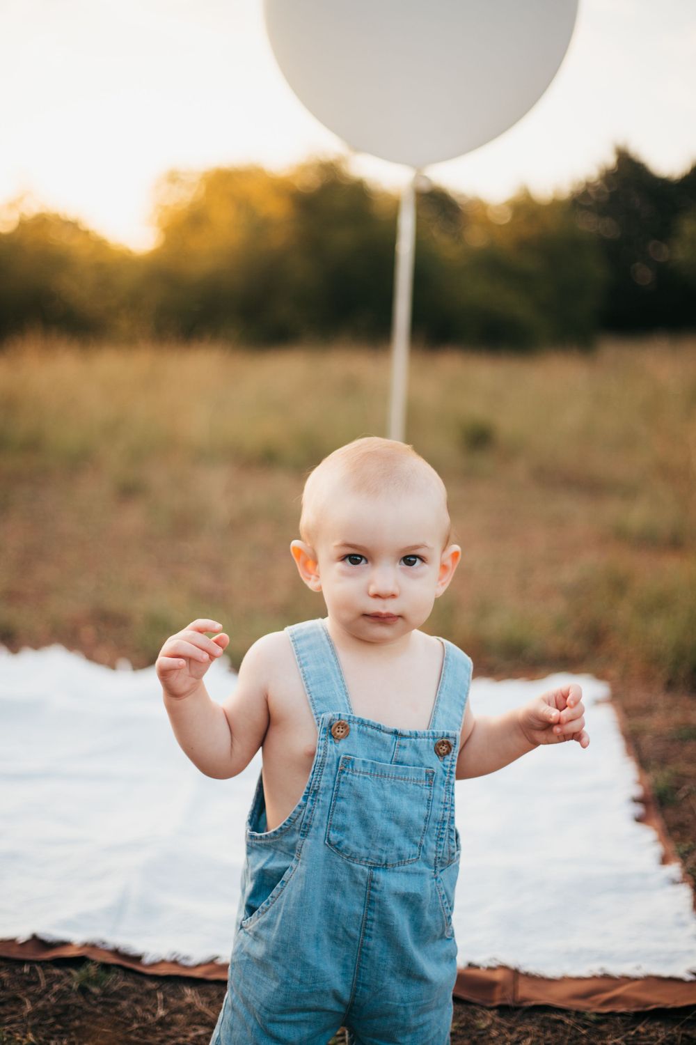 Toddler in denim overalls stands on white blanket holding large white balloon outdoors.
