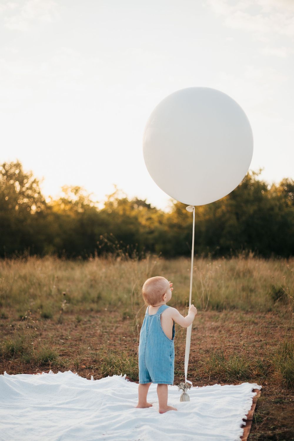 Small child in blue overalls stands on white blanket holding giant white balloon in golden field.
