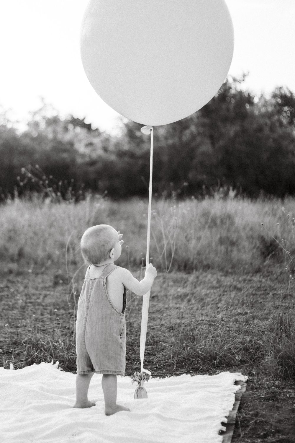 Black and white photo of toddler holding large white balloon while standing on white blanket.
