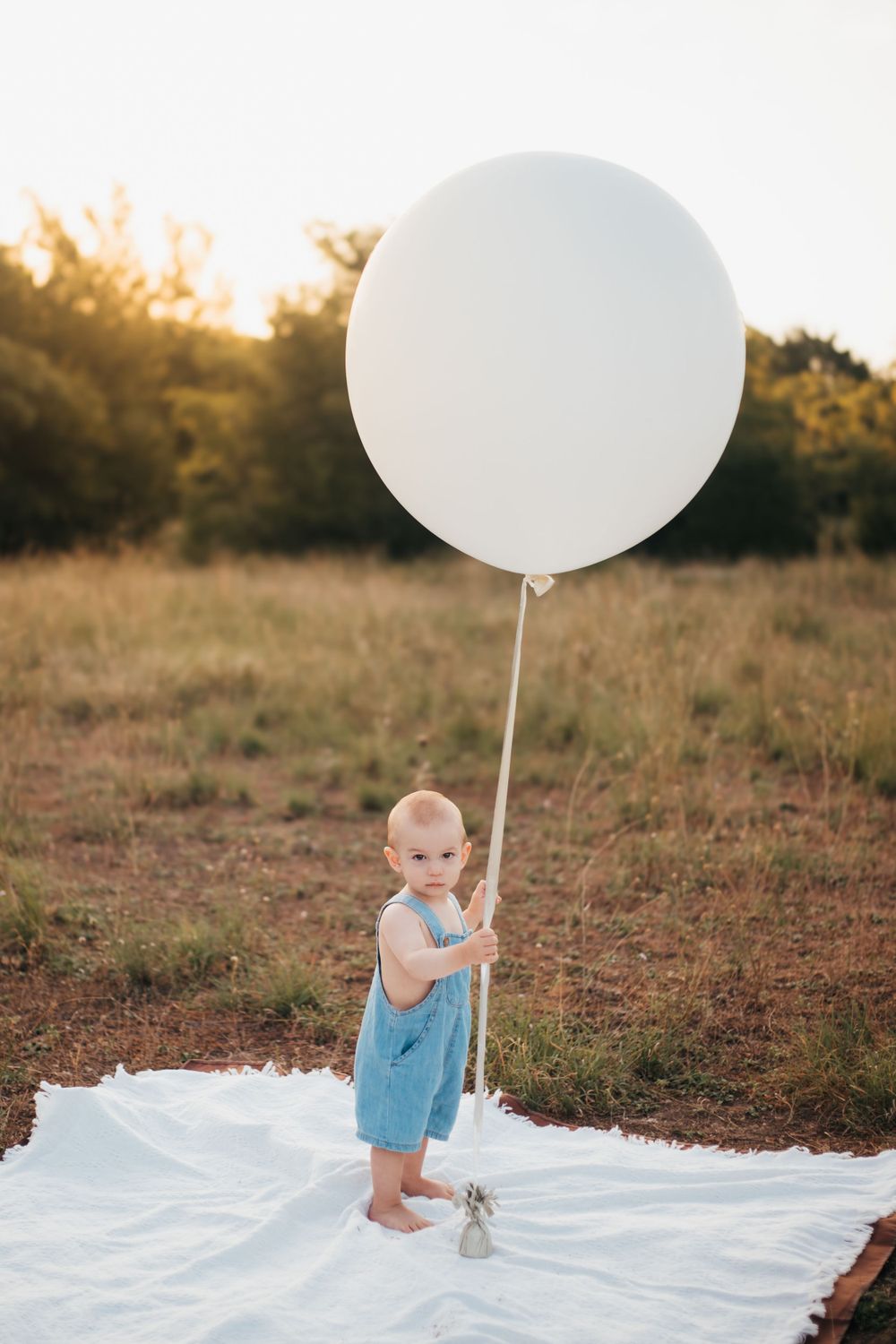 Small child in blue overalls stands barefoot on white blanket holding large white balloon at sunset.