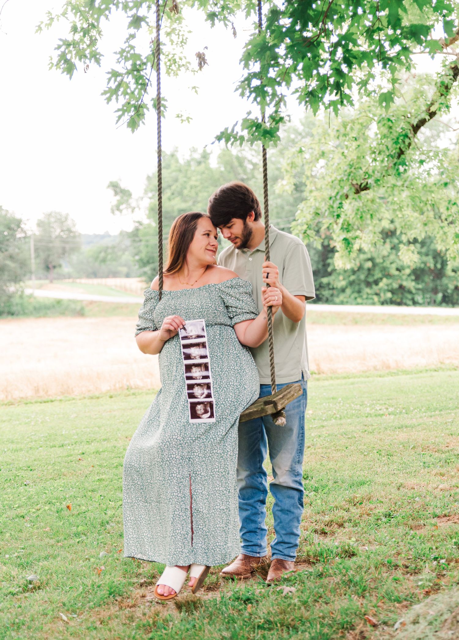 A couple sitting on a swing under an oak tree with a photo of their ultrasound of a new baby.