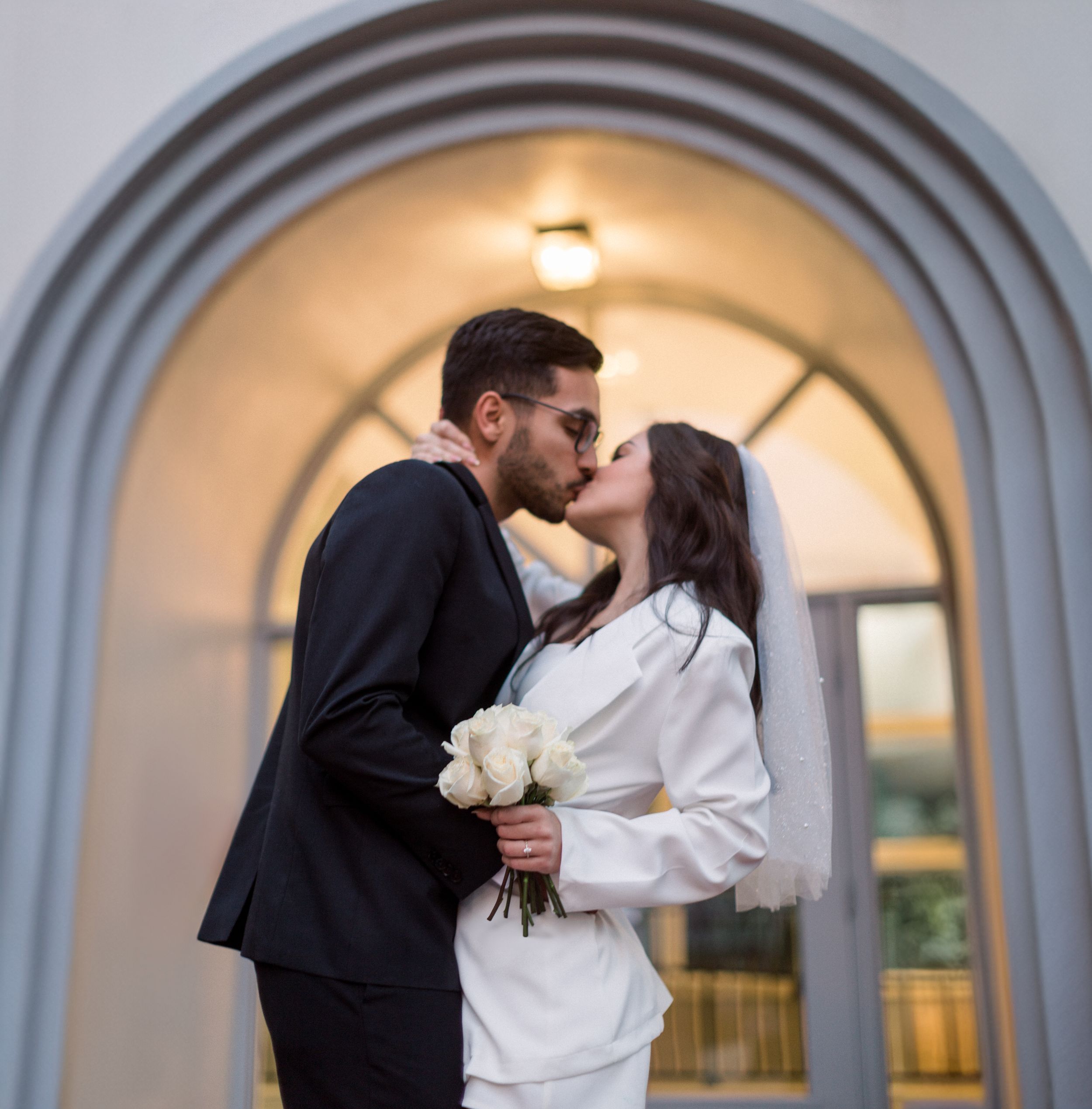 A romantic moment between newlyweds sharing a kiss under an arched doorway with white roses.