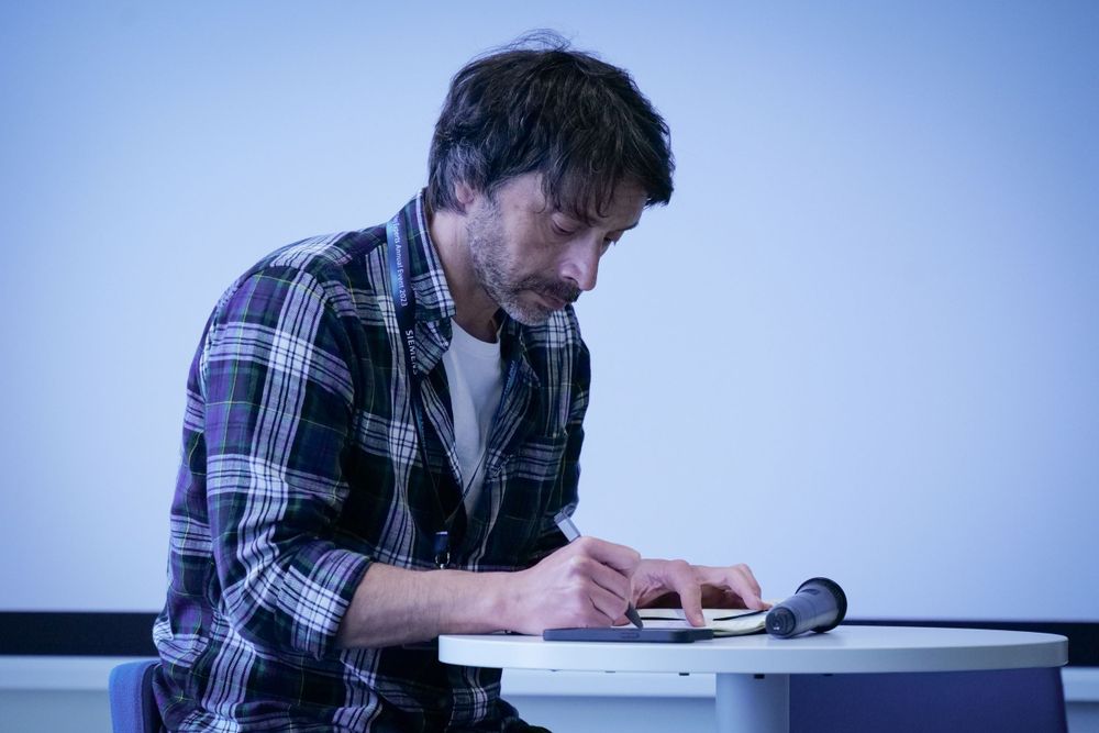 Man in plaid shirt writing notes at a desk during a conference or seminar.