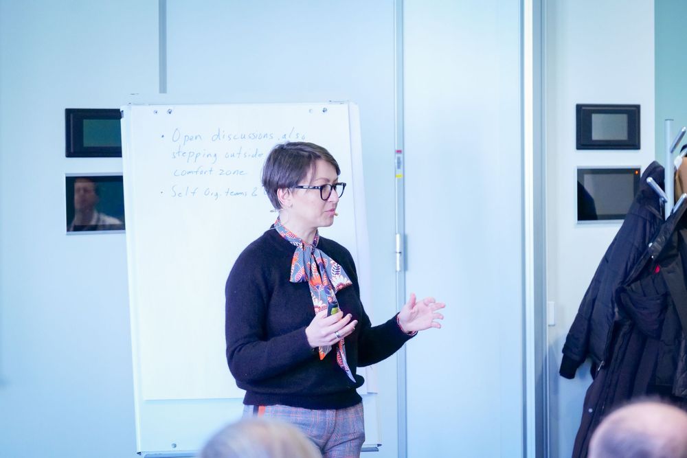 Woman in black sweater presenting at a business workshop near a whiteboard.