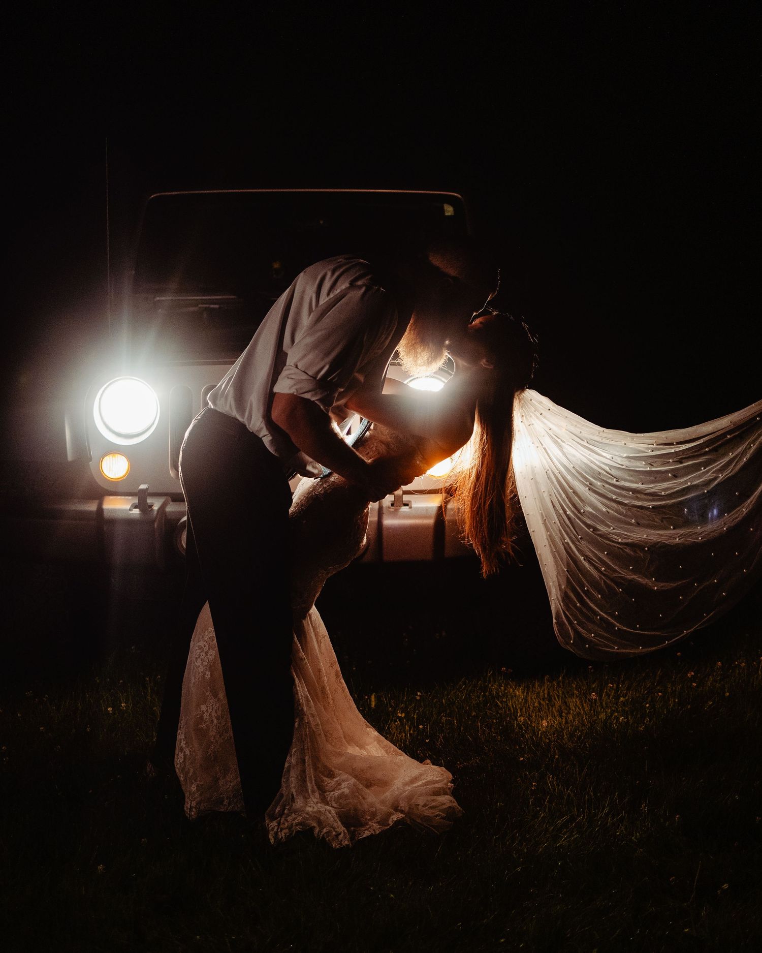 Dramatic nighttime wedding photo with vintage car headlights illuminating the couple.
