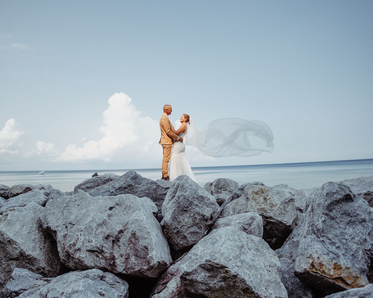 A couple stands on rocky coastal outcrop with ocean and cloudy sky in background.