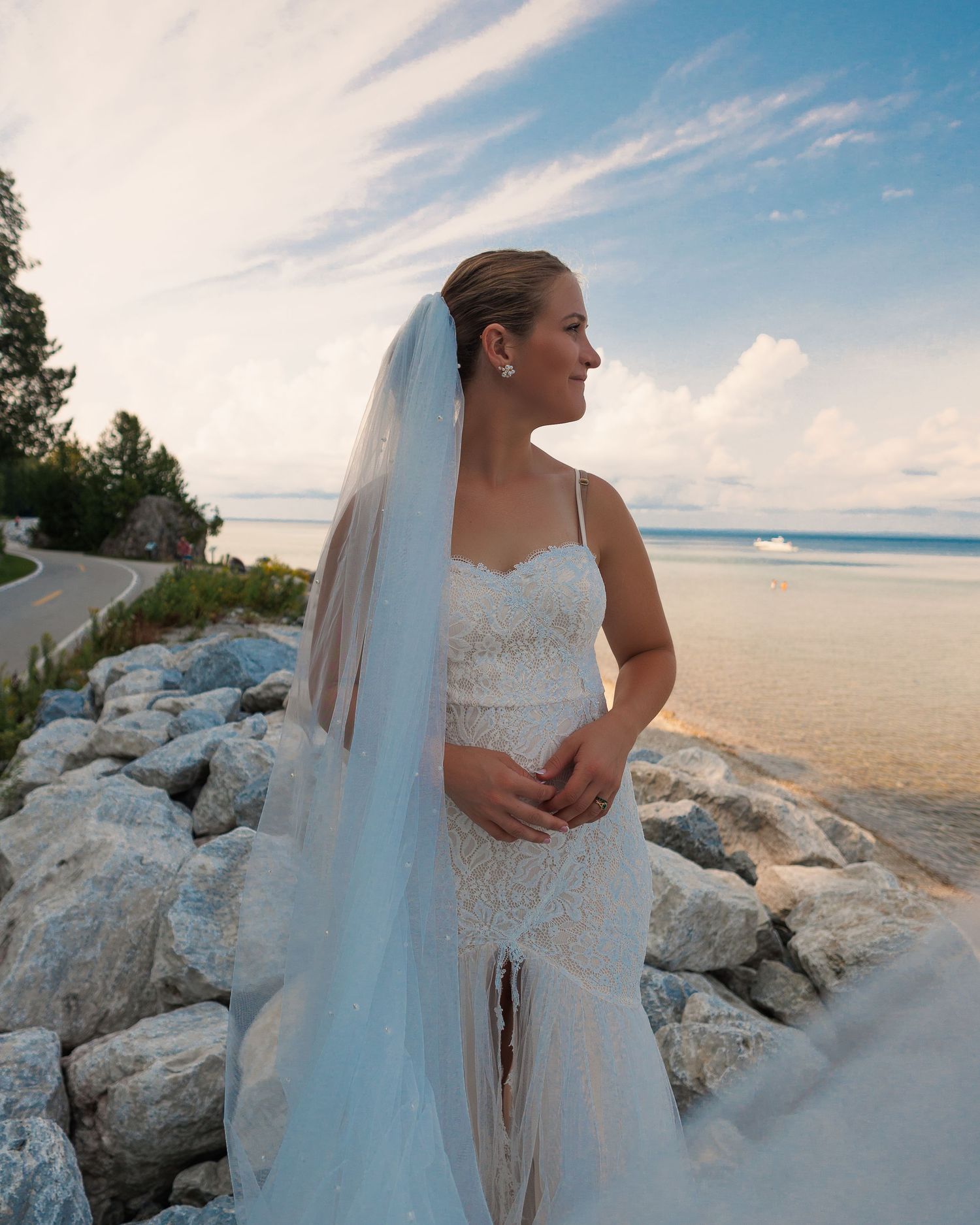 Bride in lace dress standing on rocky beach overlook during sunset.