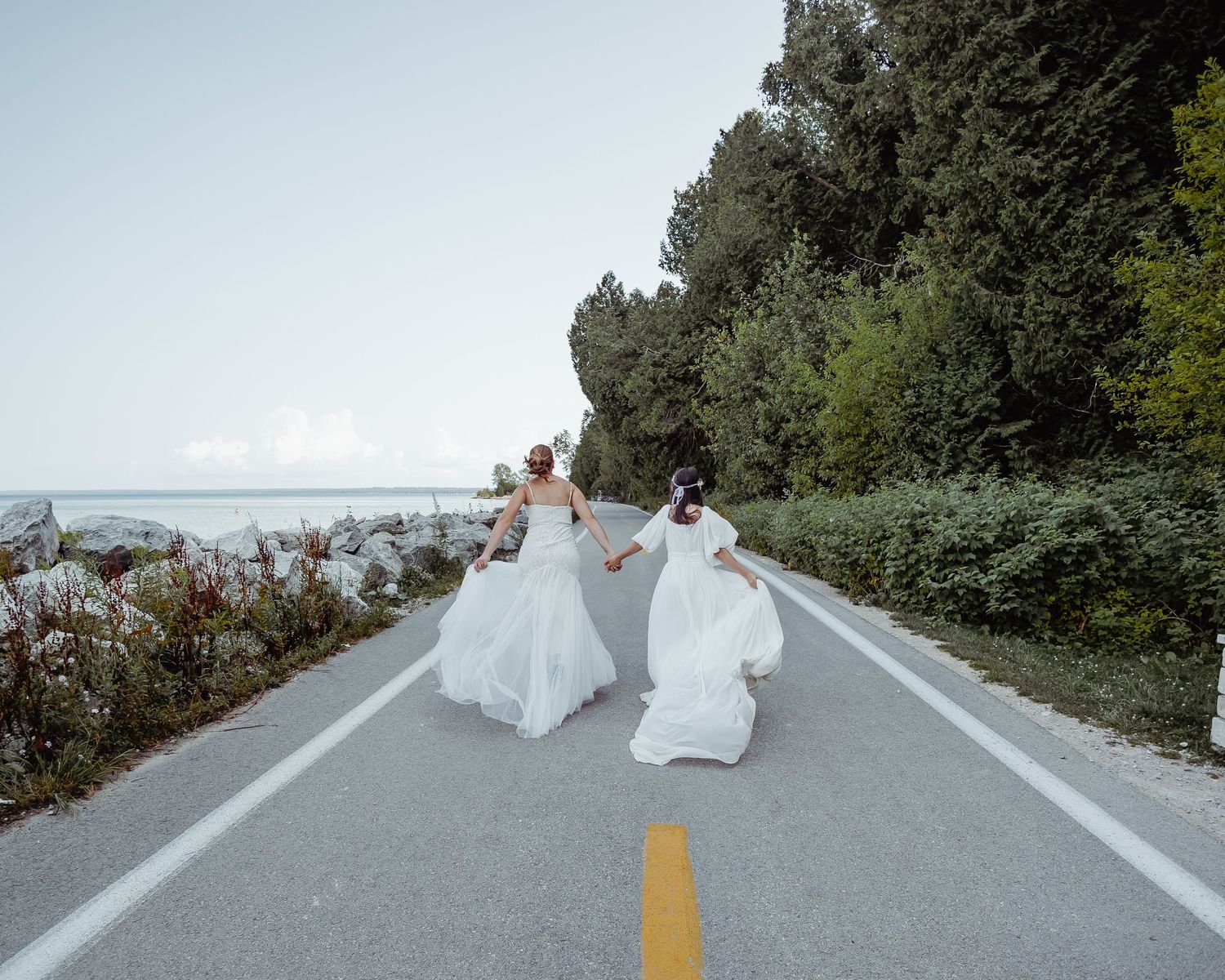 Two people in wedding dresses running down an empty coastal road.