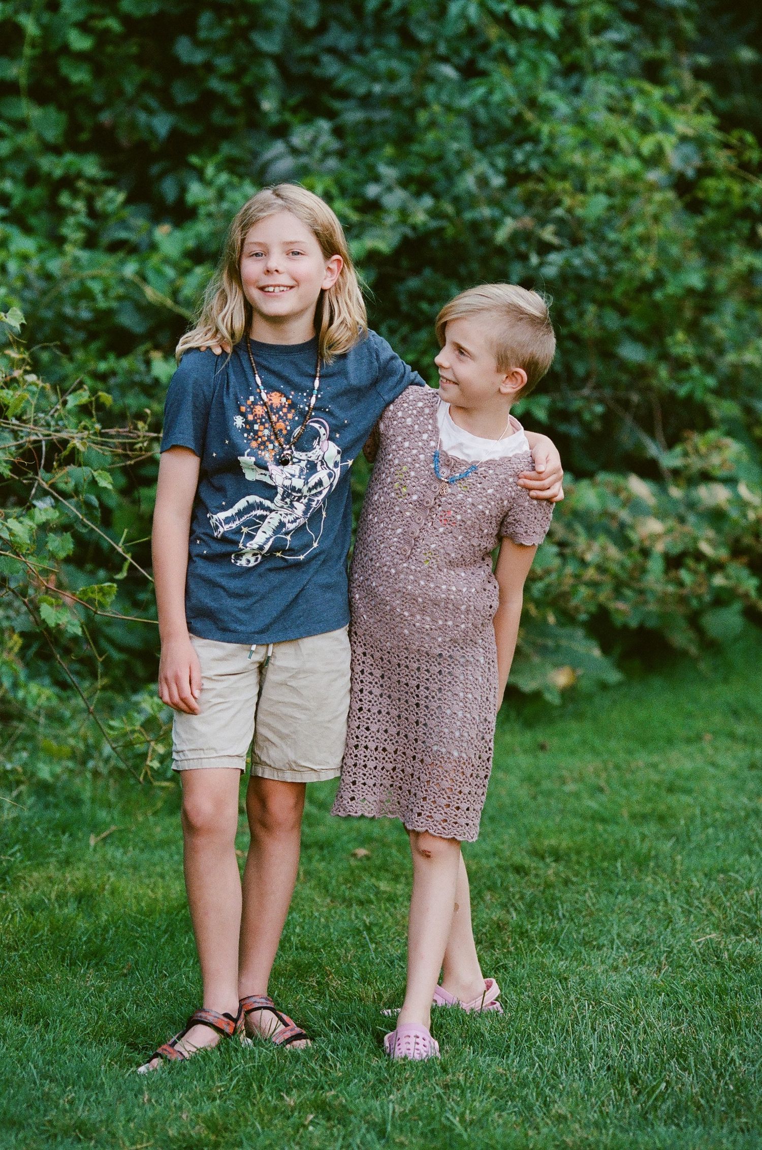 Two children standing together on a lush green lawn with trees in background.