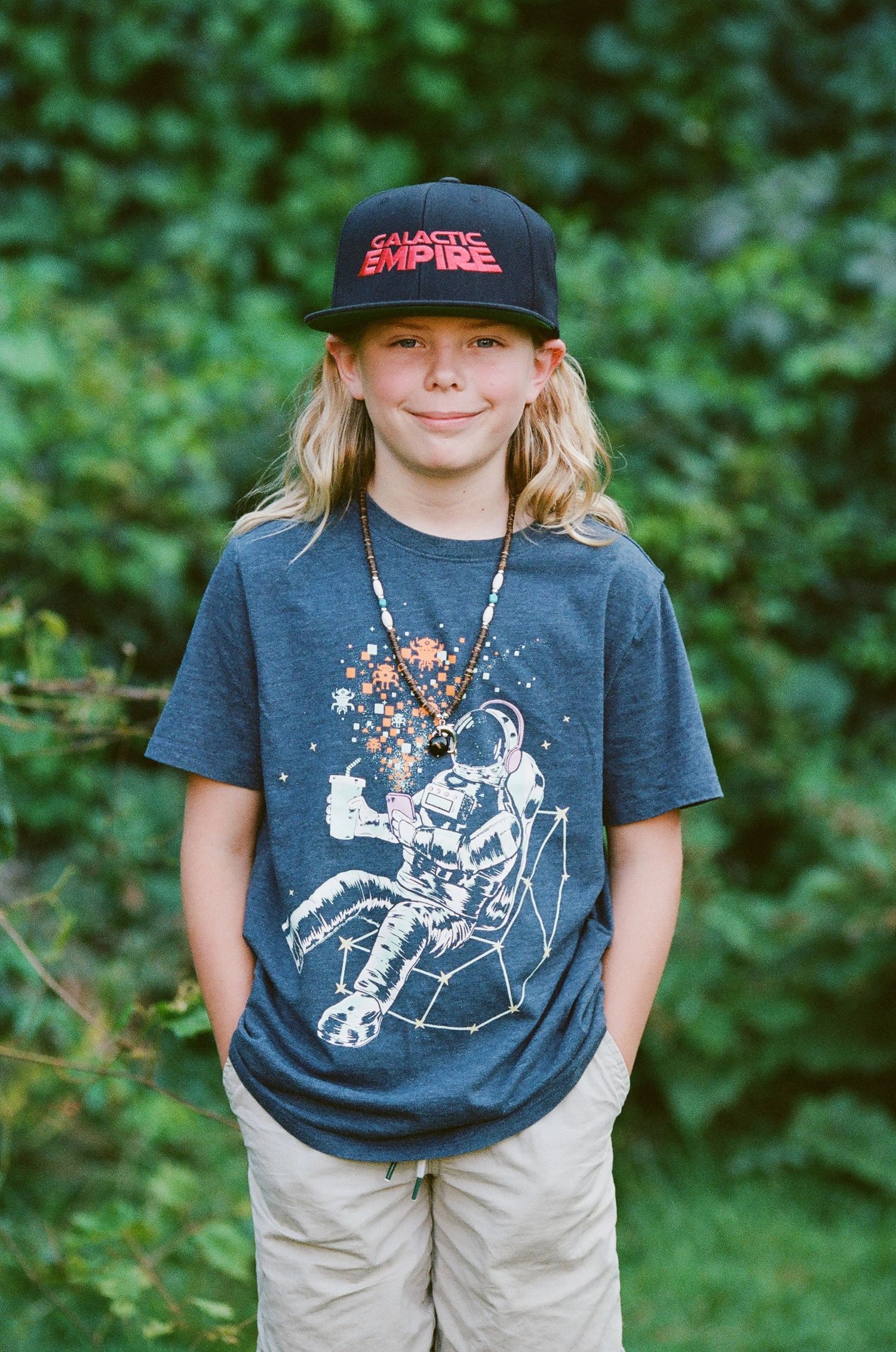 Child wearing baseball cap and graphic t-shirt standing outdoors.