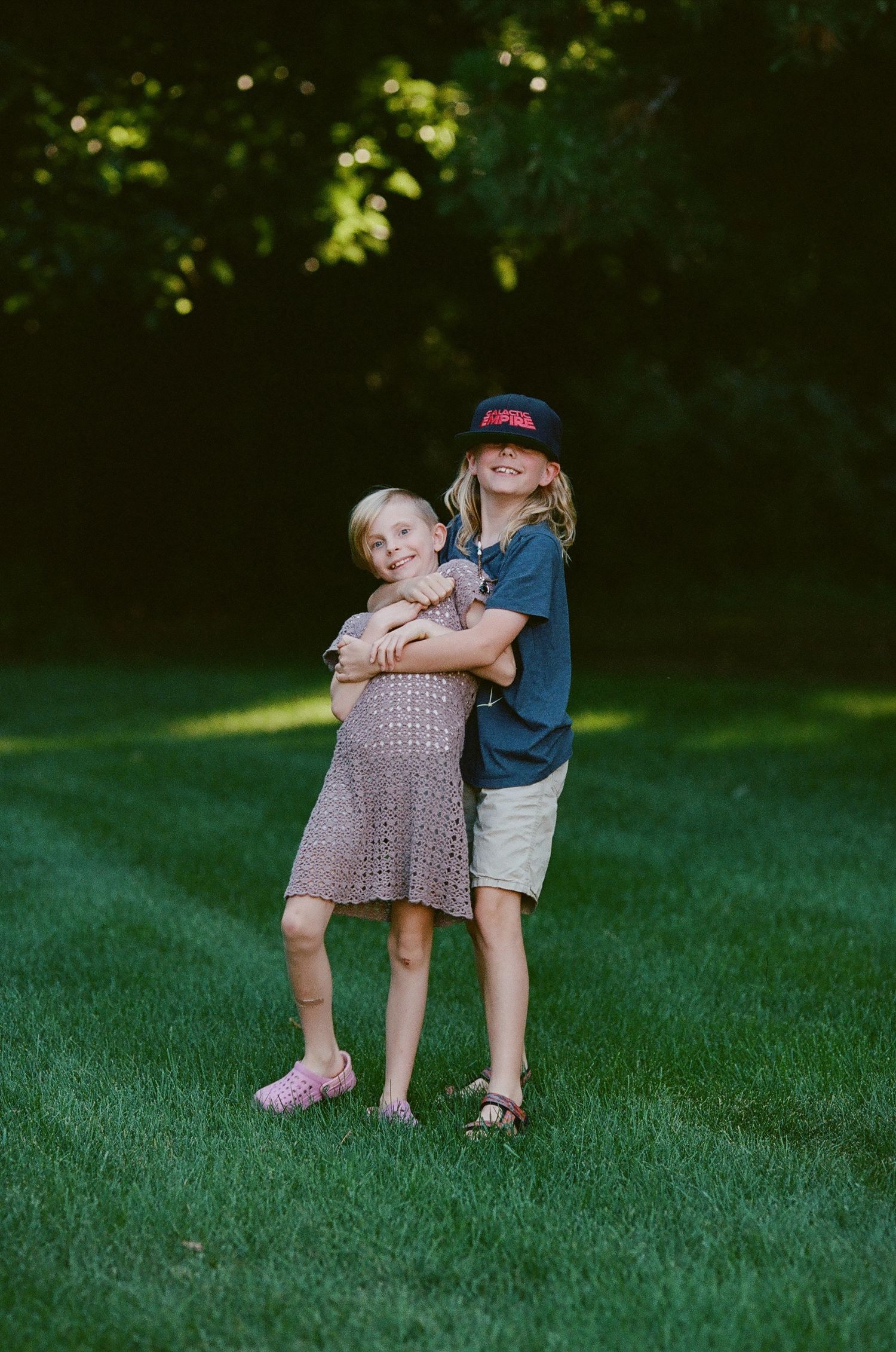Two children embracing on a sunlit grass lawn with trees in background.