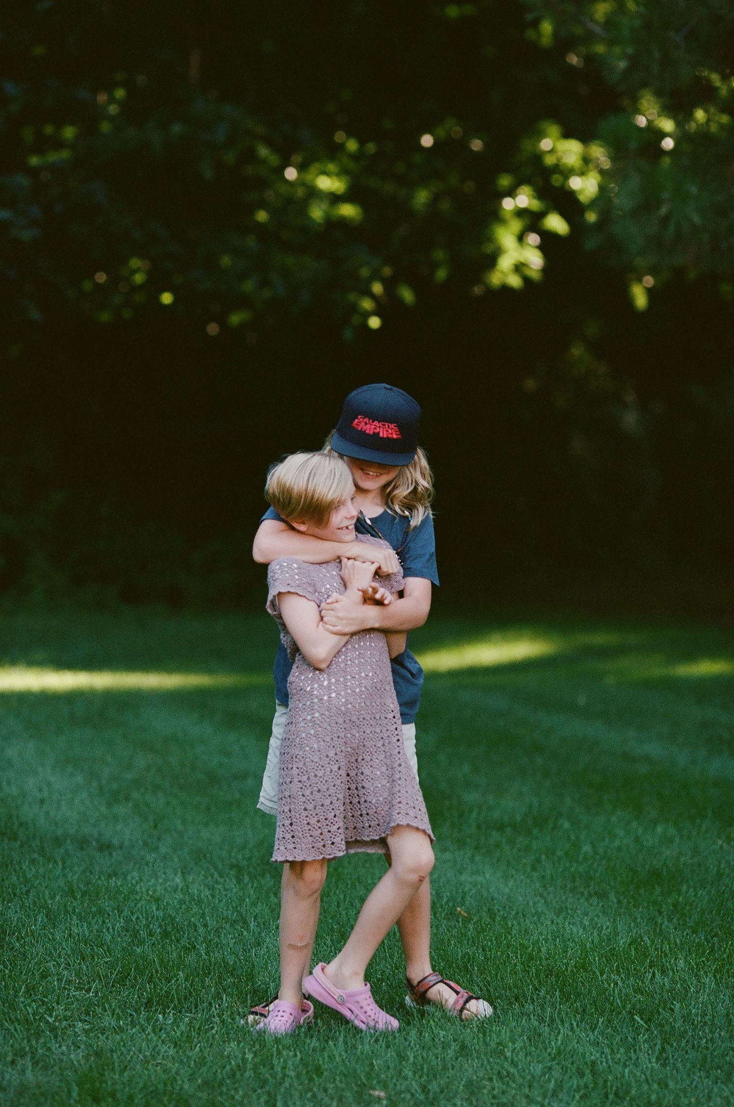 Two children hugging on a sunny lawn with green foliage in background.