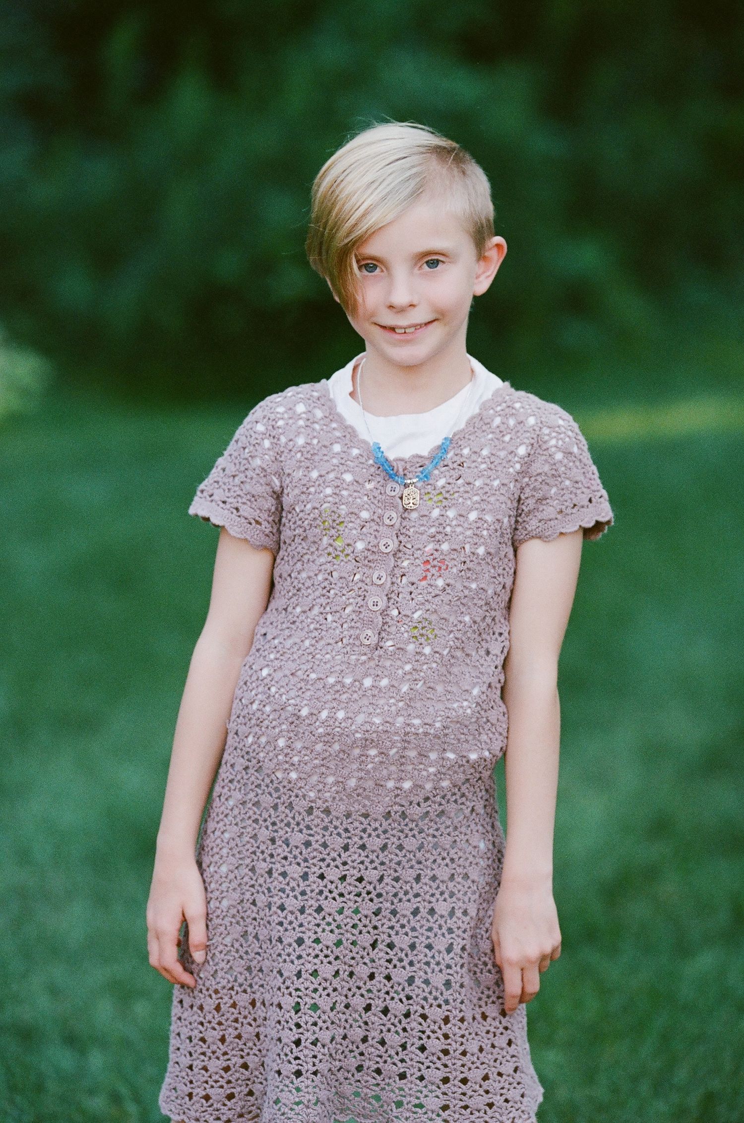 A child wearing a brown crochet dress stands outdoors in a garden setting.