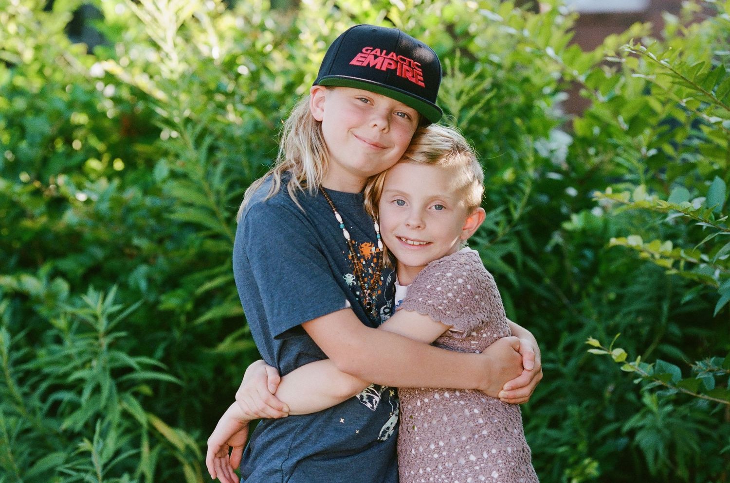 Two children share a warm embrace while standing in front of green foliage.