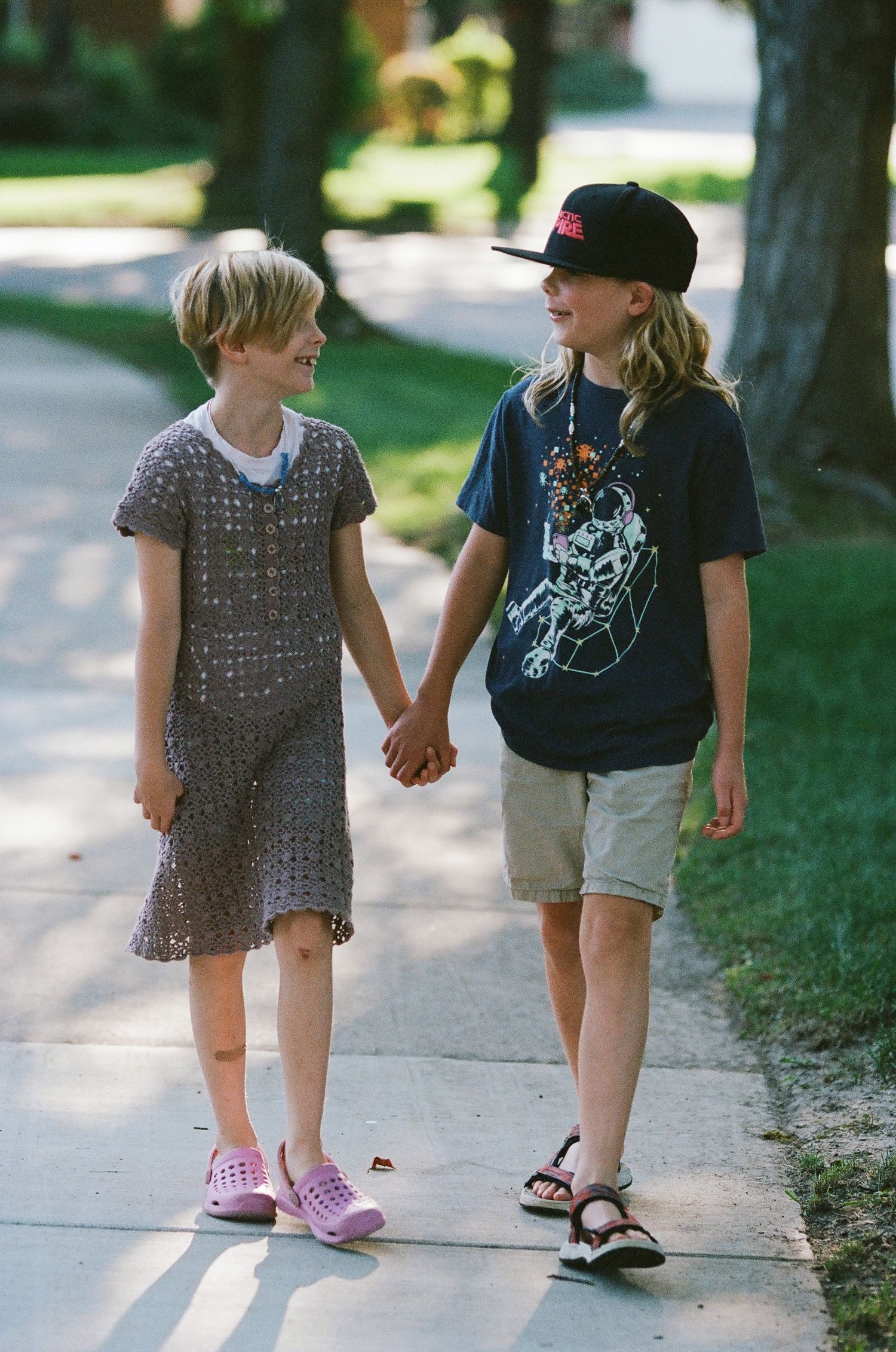 Two children holding hands while walking down a neighborhood sidewalk.