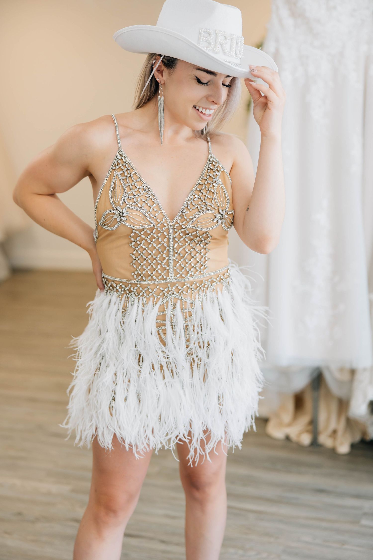 Smiling bride in embellished feather mini dress tipping white BRIDE cowboy hat in boutique.