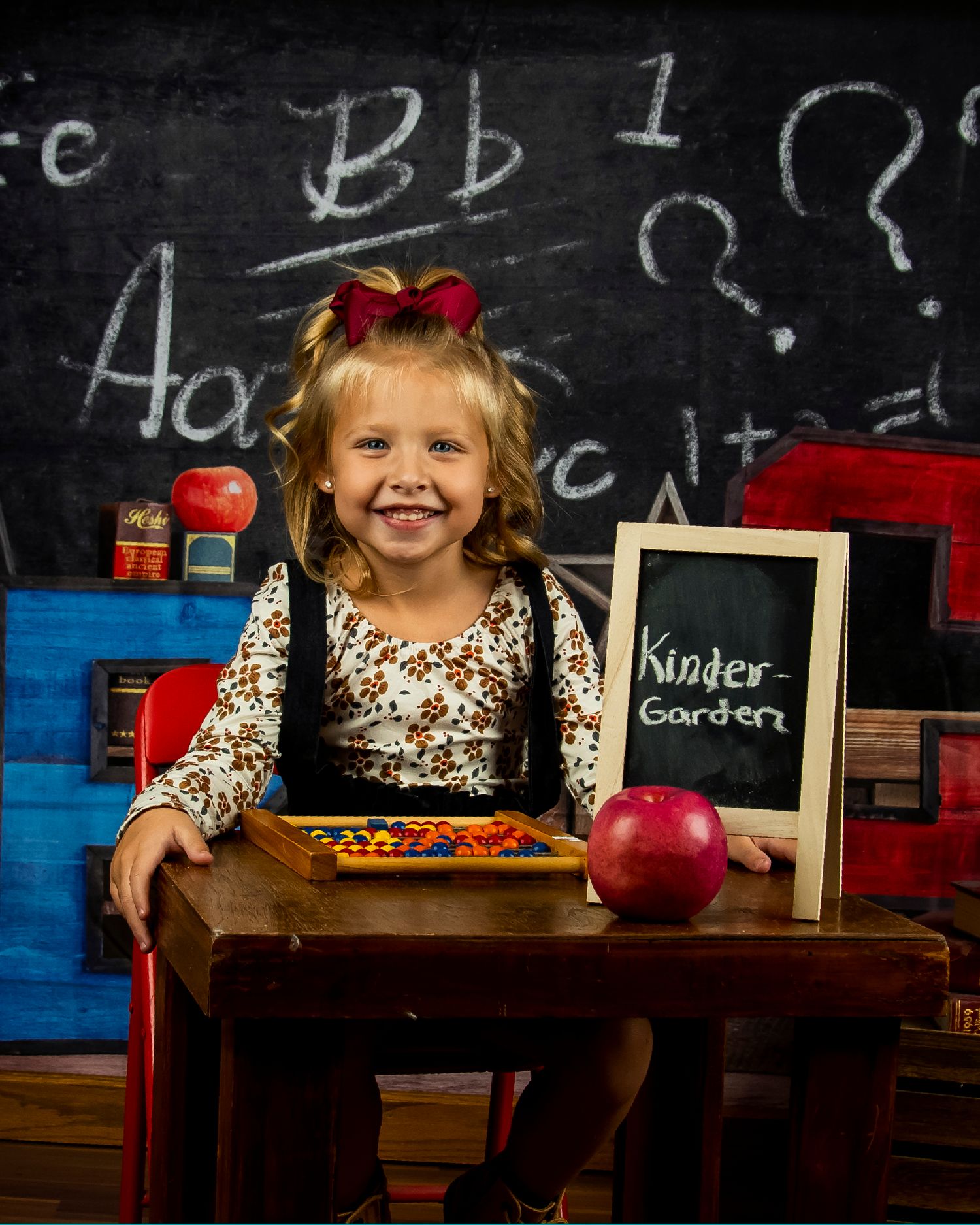 A school-themed portrait setup with a chalkboard background, small desk, red apple, and kindergarten sign in a classroom setting.