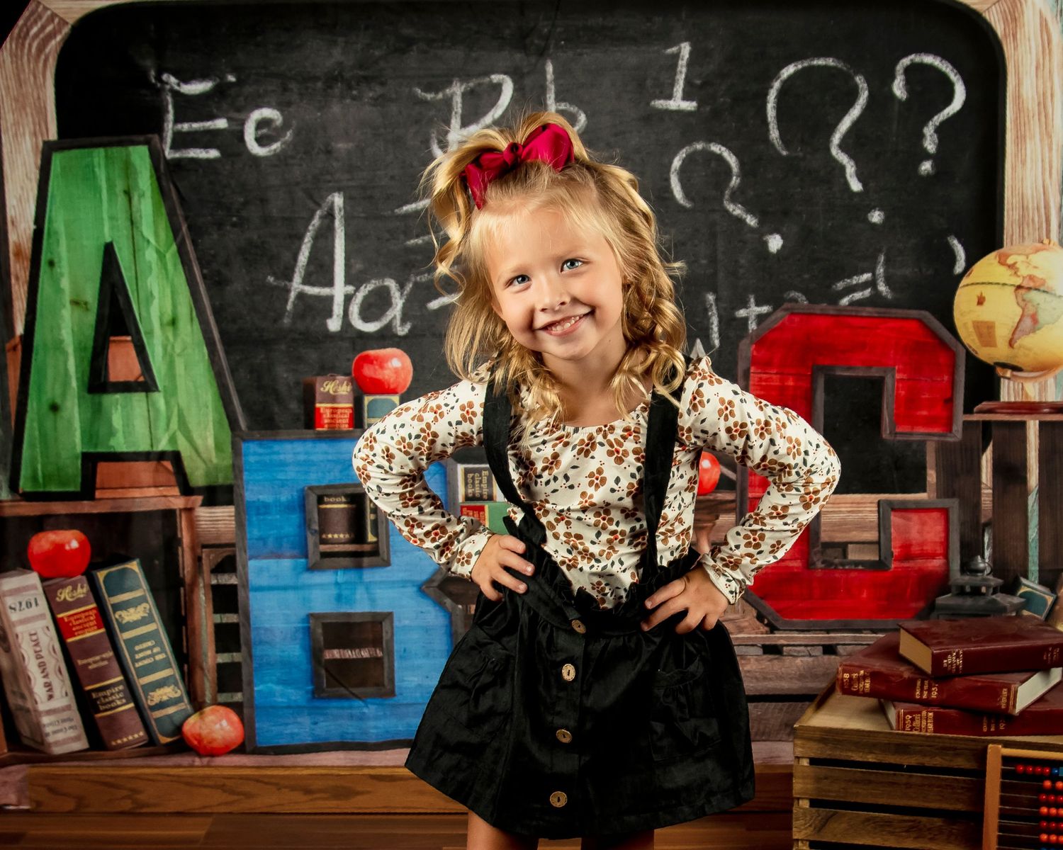 A colorful classroom scene with ABC blocks, books, and apples on a desk in front of a chalkboard with question marks.