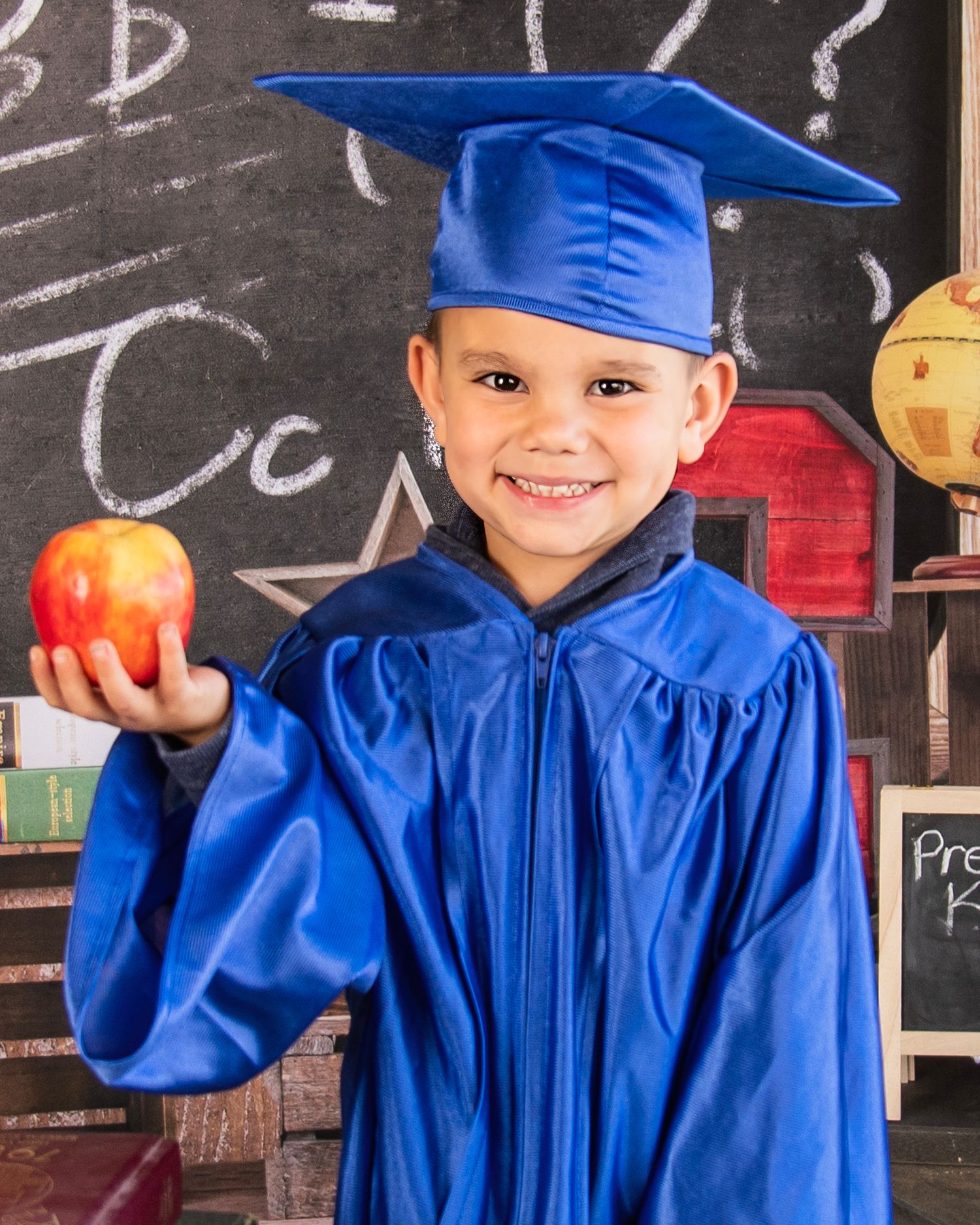 Graduation portrait in blue cap and gown holding red apple against chalkboard.