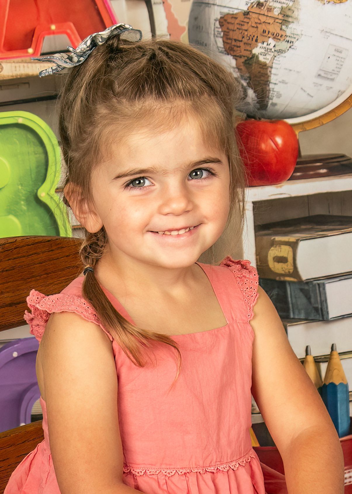 A young child in a coral pink dress sits smiling in front of colorful toys and educational materials in what appears to be a playroom.