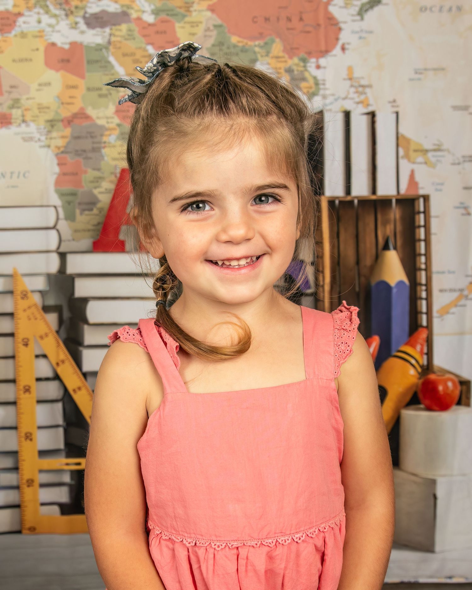 A child wearing a coral pink dress poses for a school photo in front of a world map and educational supplies.