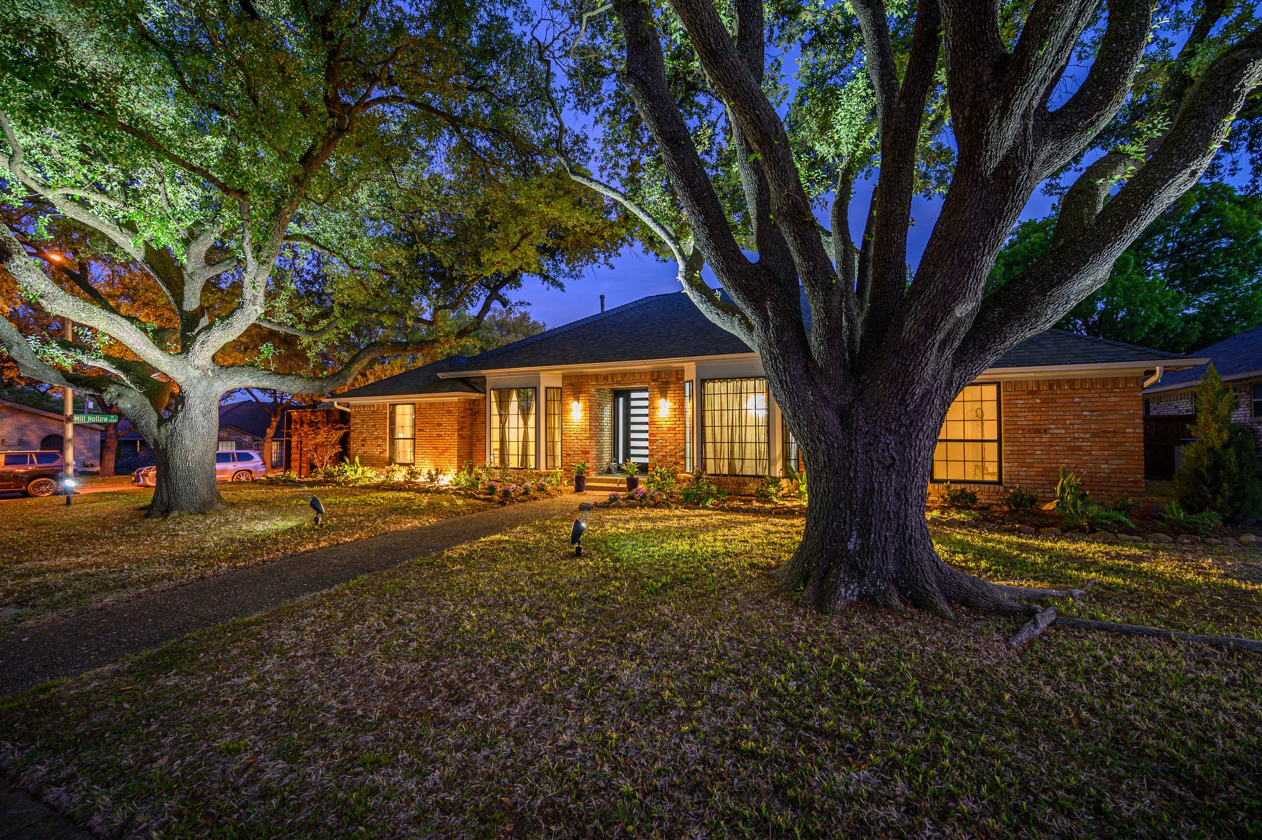 Ranch-style home illuminated at dusk with large oak trees casting shadows across the front yard.