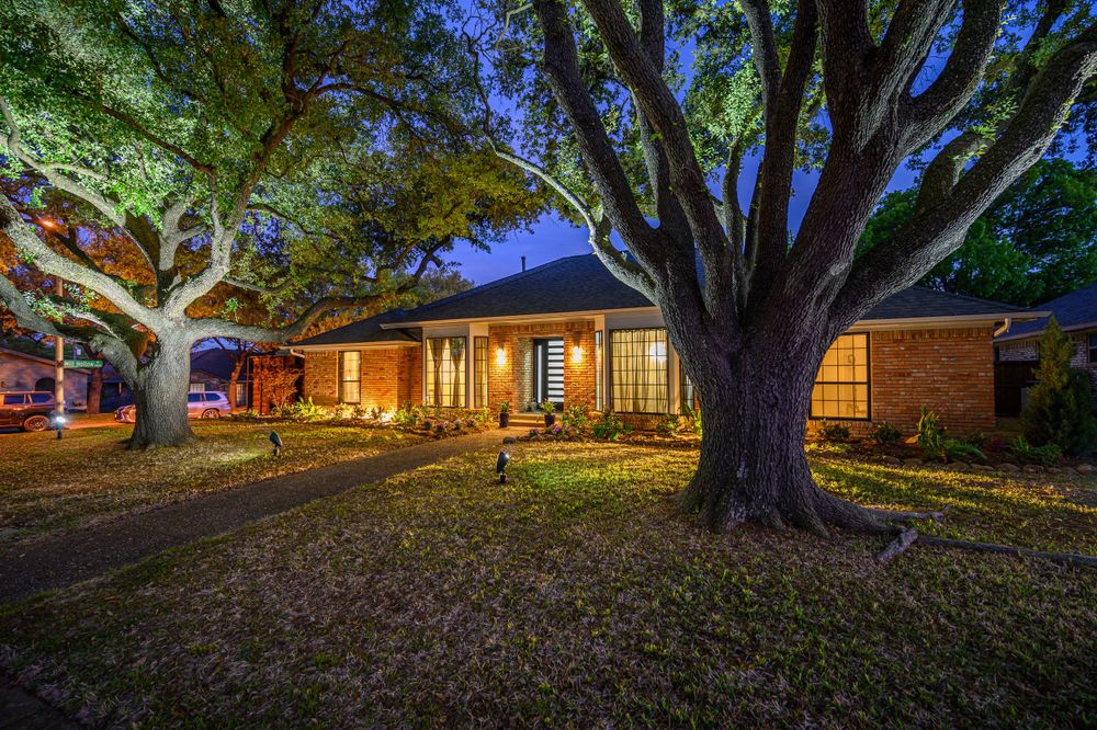 Ranch-style home illuminated at dusk with large oak trees casting shadows across the front yard.
