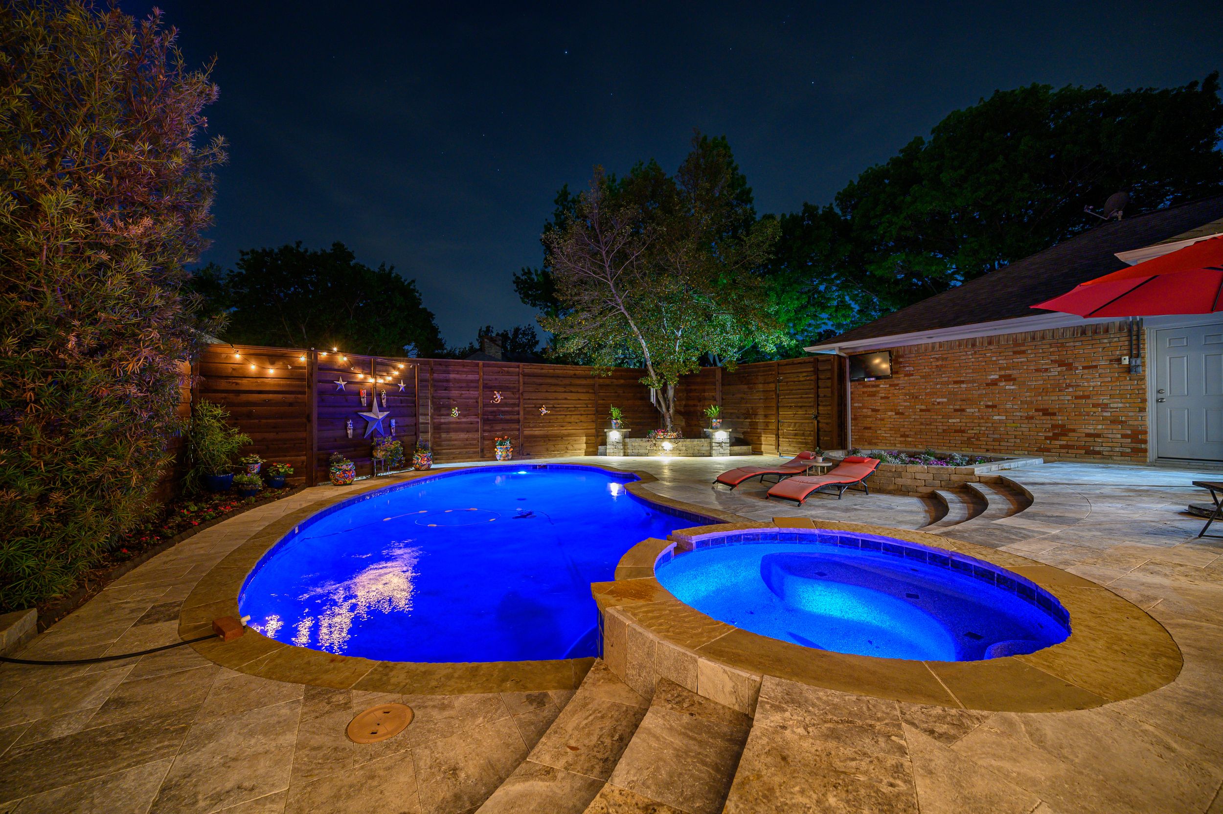Illuminated backyard featuring a curved blue swimming pool and spa with string lights overhead at night.