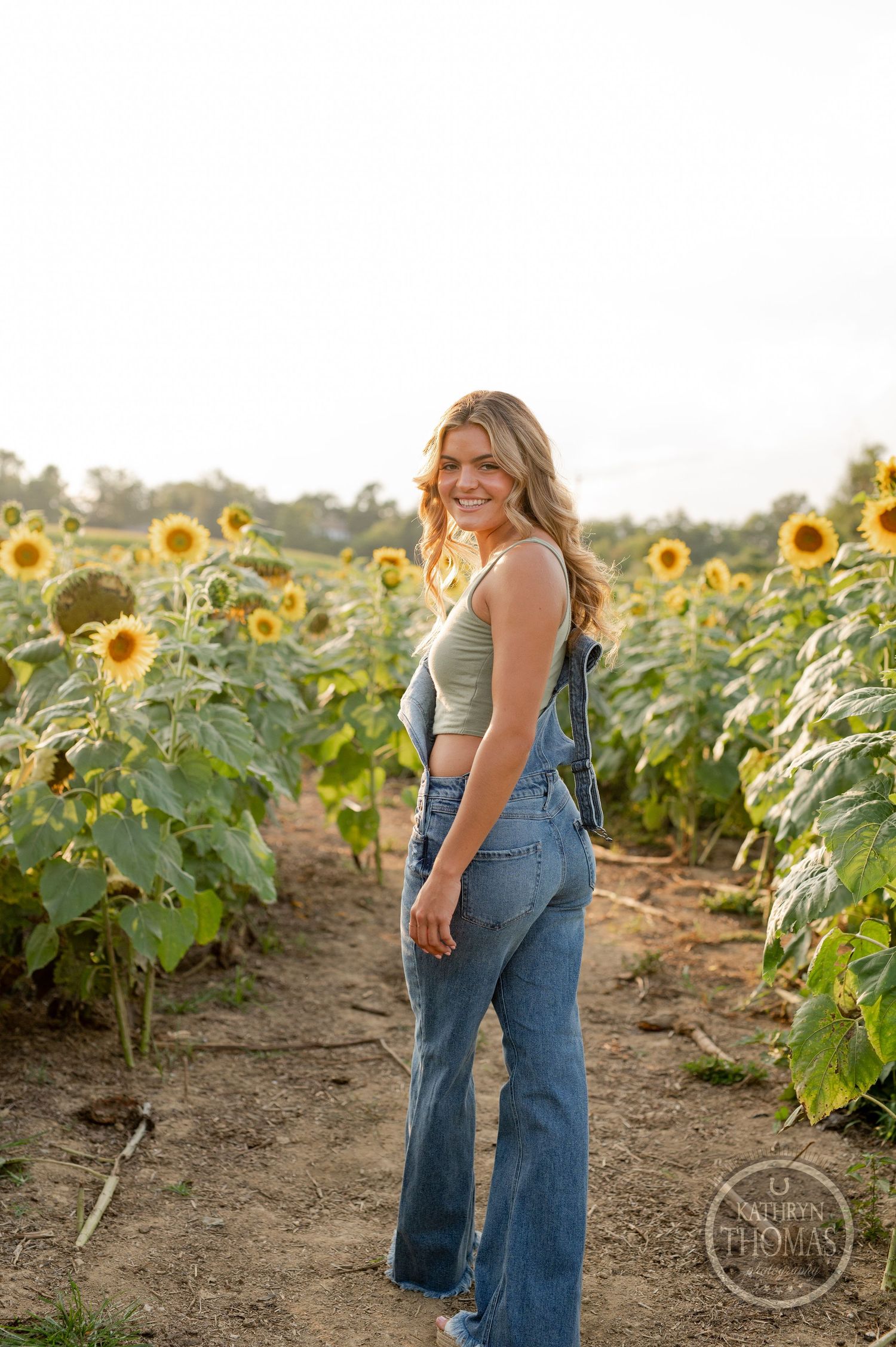 A person in denim overalls and tank top stands in a vibrant sunflower field during golden hour.