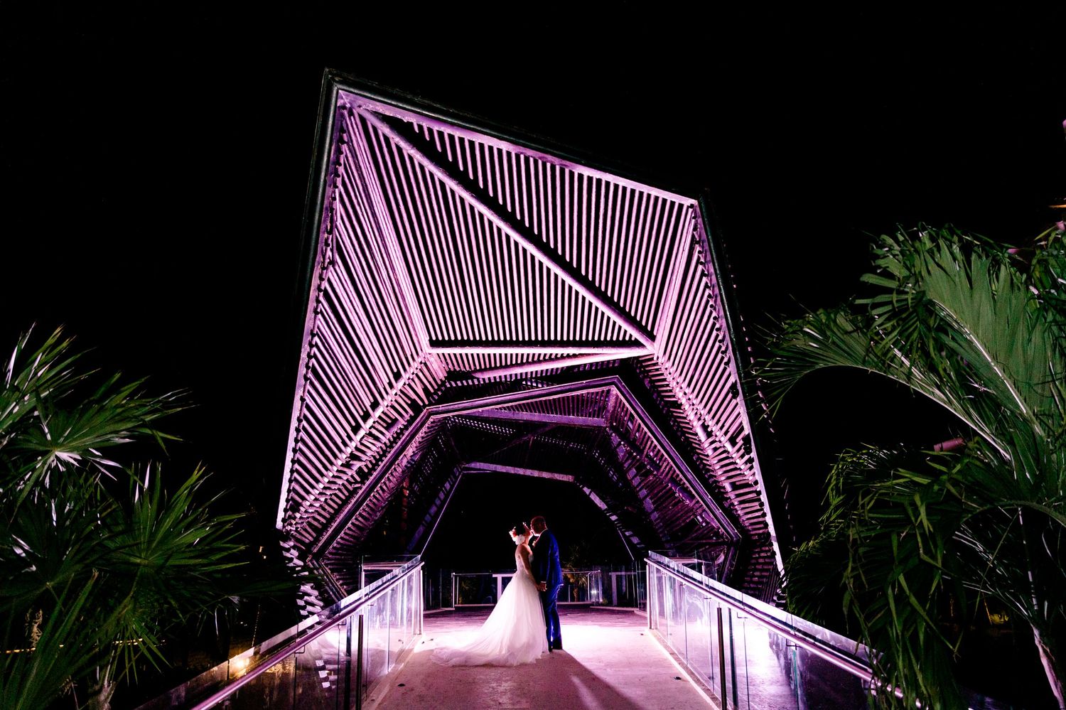 Illuminated purple geometric bridge walkway creates dramatic wedding photo backdrop at night with tropical foliage.