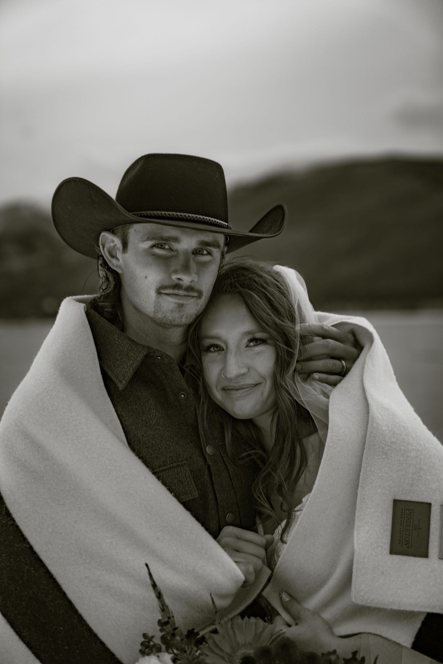 A bride and groom embrace, wrapped in a white blanket while wearing western attire in a black and white photo.