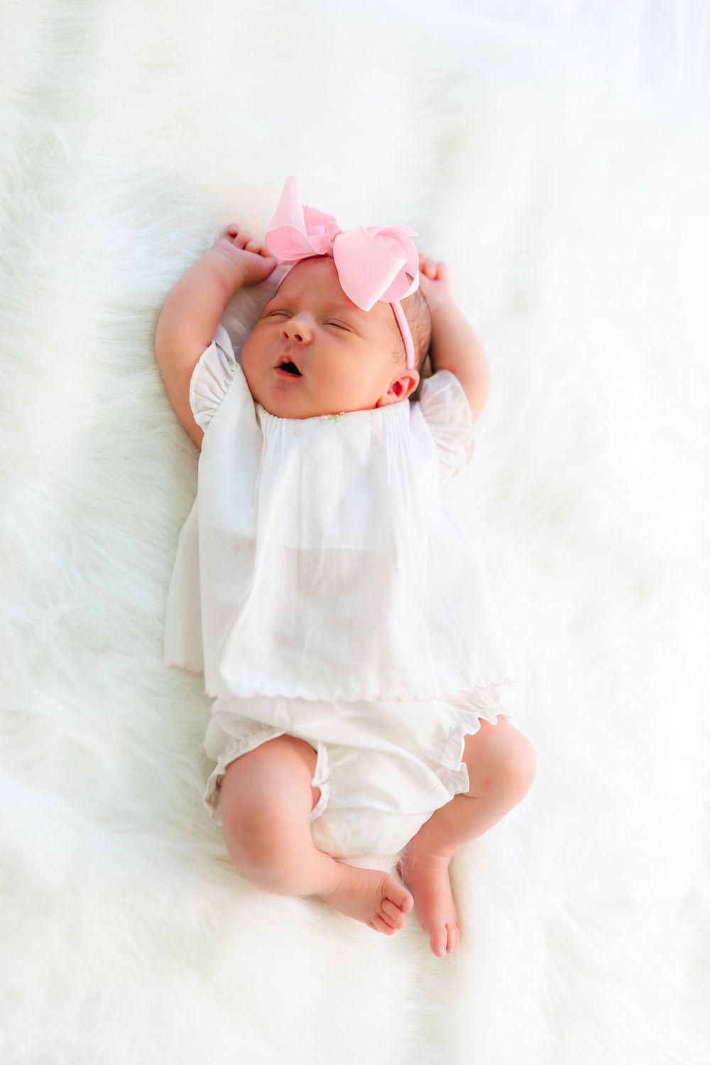 Newborn baby wearing white outfit and pink bow lying on soft white blanket.