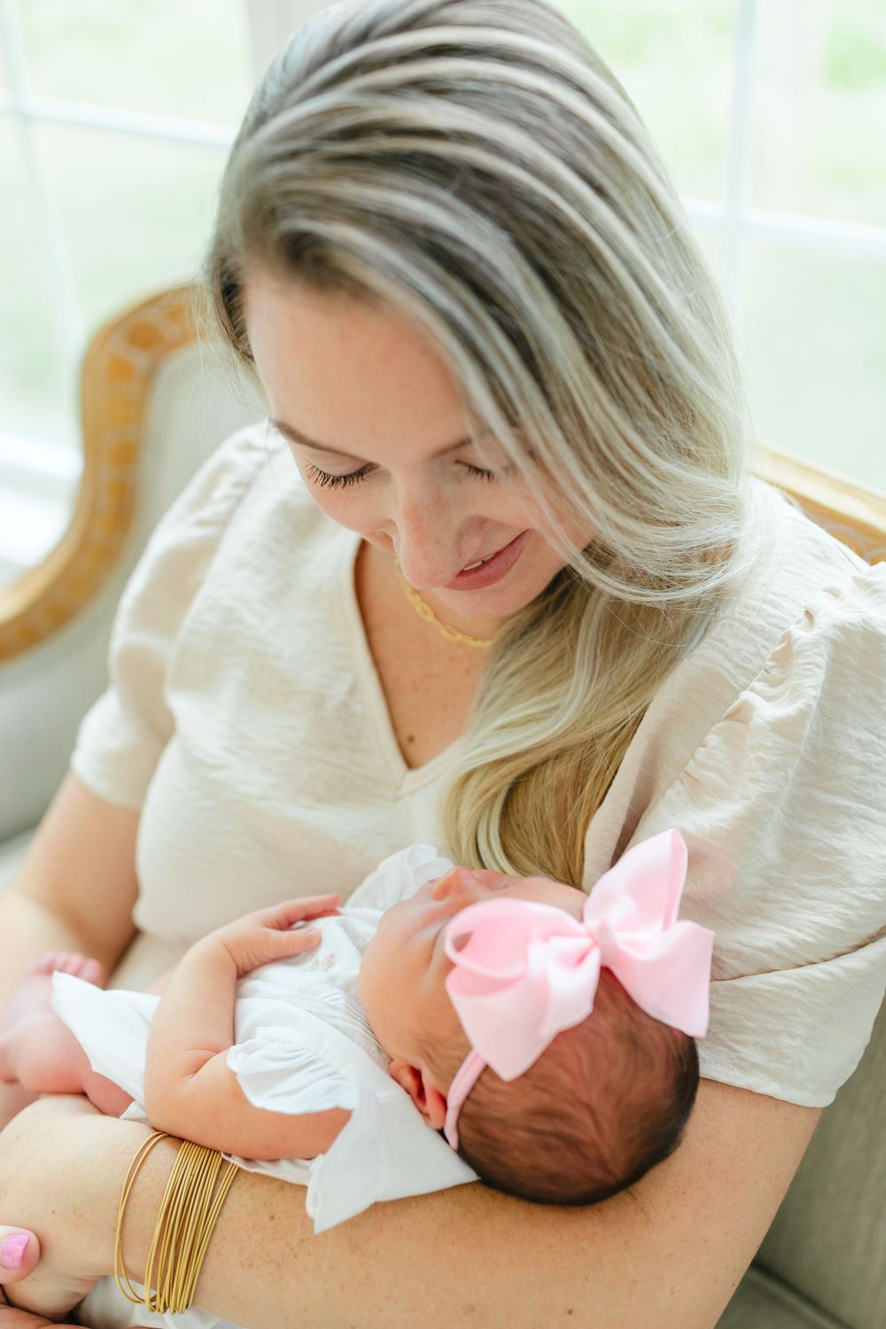 Mother in white dress tenderly holding newborn baby with pink bow.