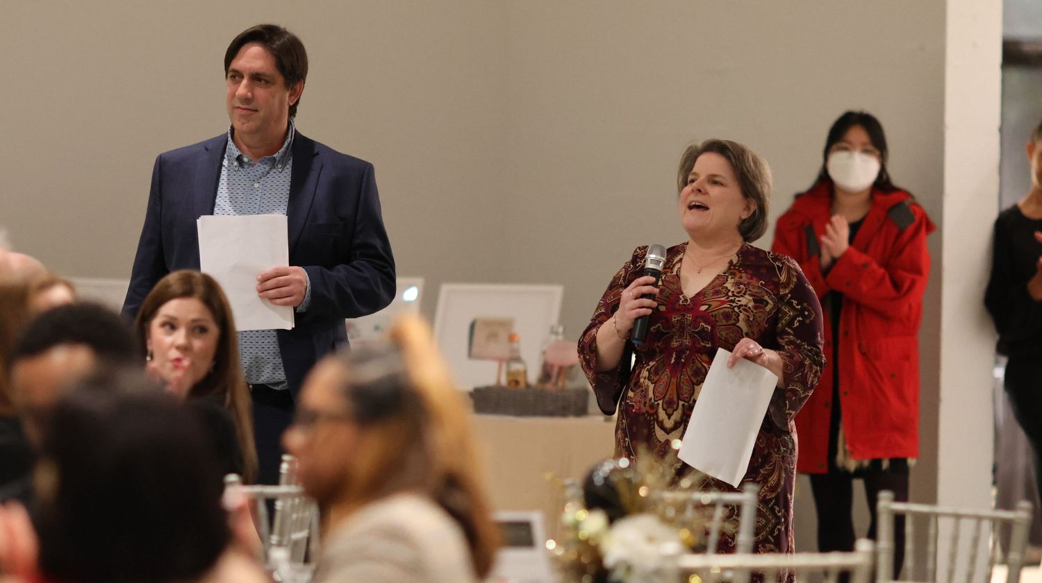 People speaking at a formal dinner event with tables and chairs in the background.