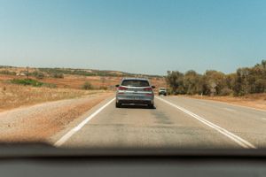 Silver car driving on empty rural road through arid countryside under clear blue sky.