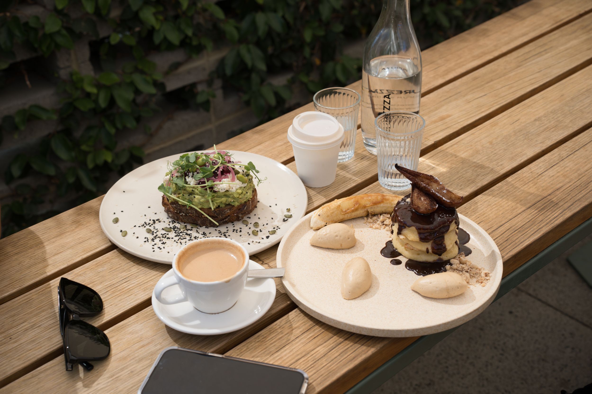Coffee and gourmet plates served on wooden table outdoors with water bottle and phone.