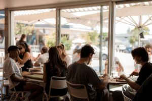 Interior view of a busy cafe with customers dining at tables near large windows.