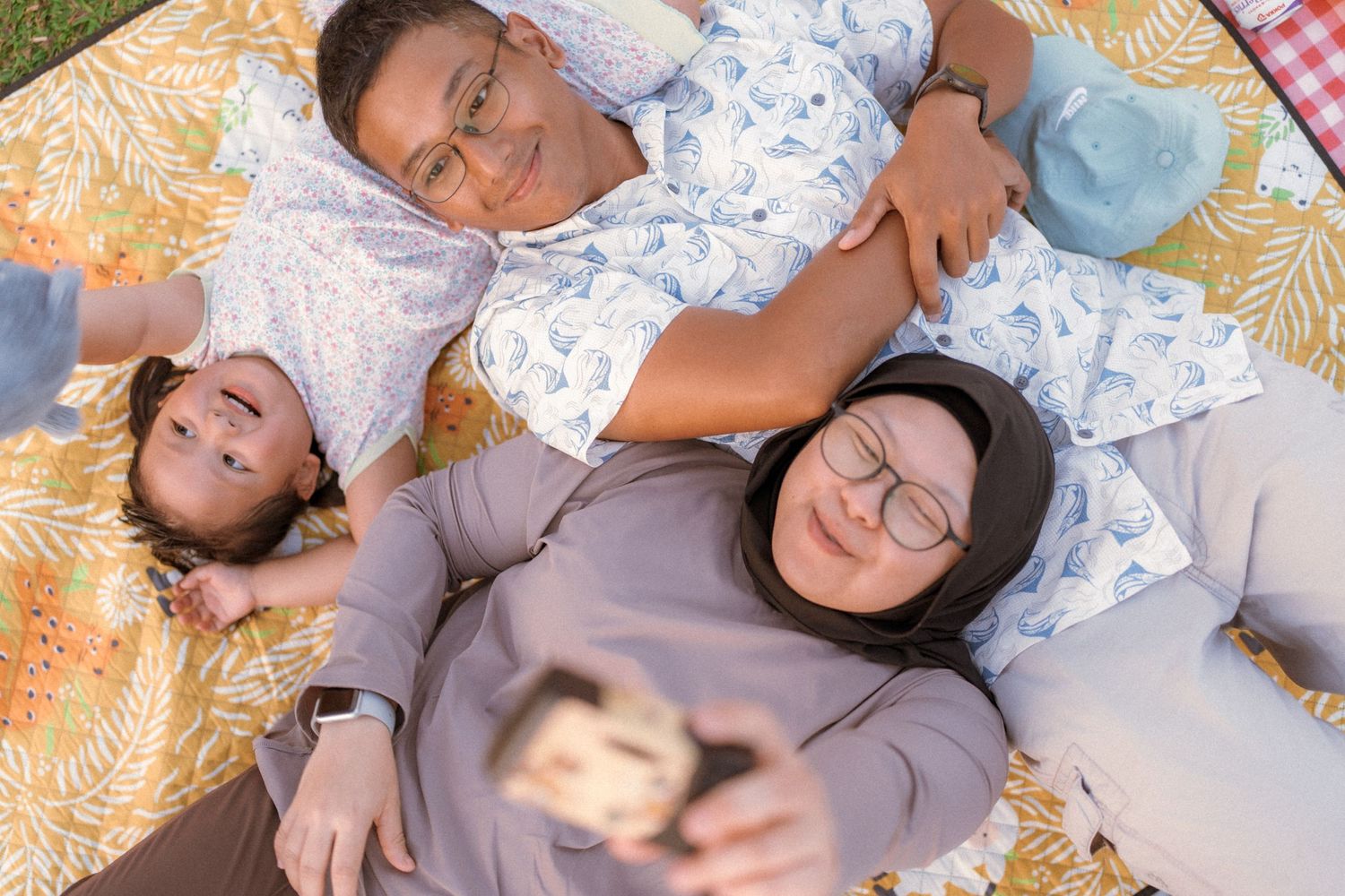 Family lying on patterned blanket taking selfie outdoors.