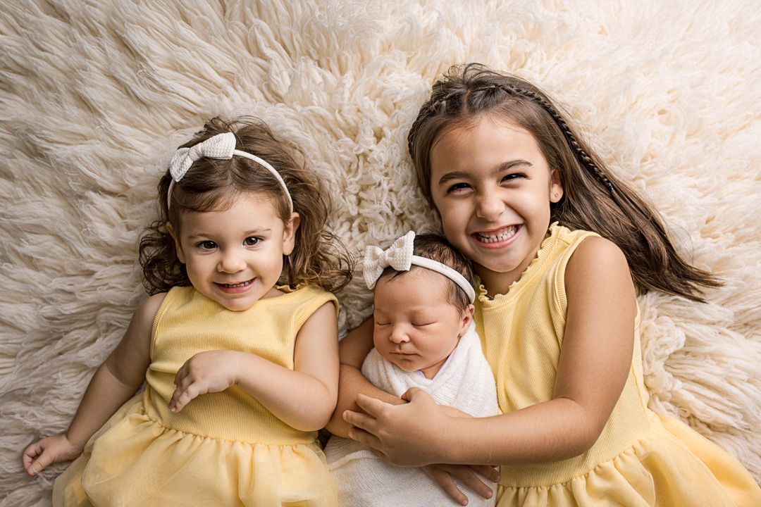 Three siblings wearing yellow dresses pose together on a fluffy cream-colored backdrop for a portrait.