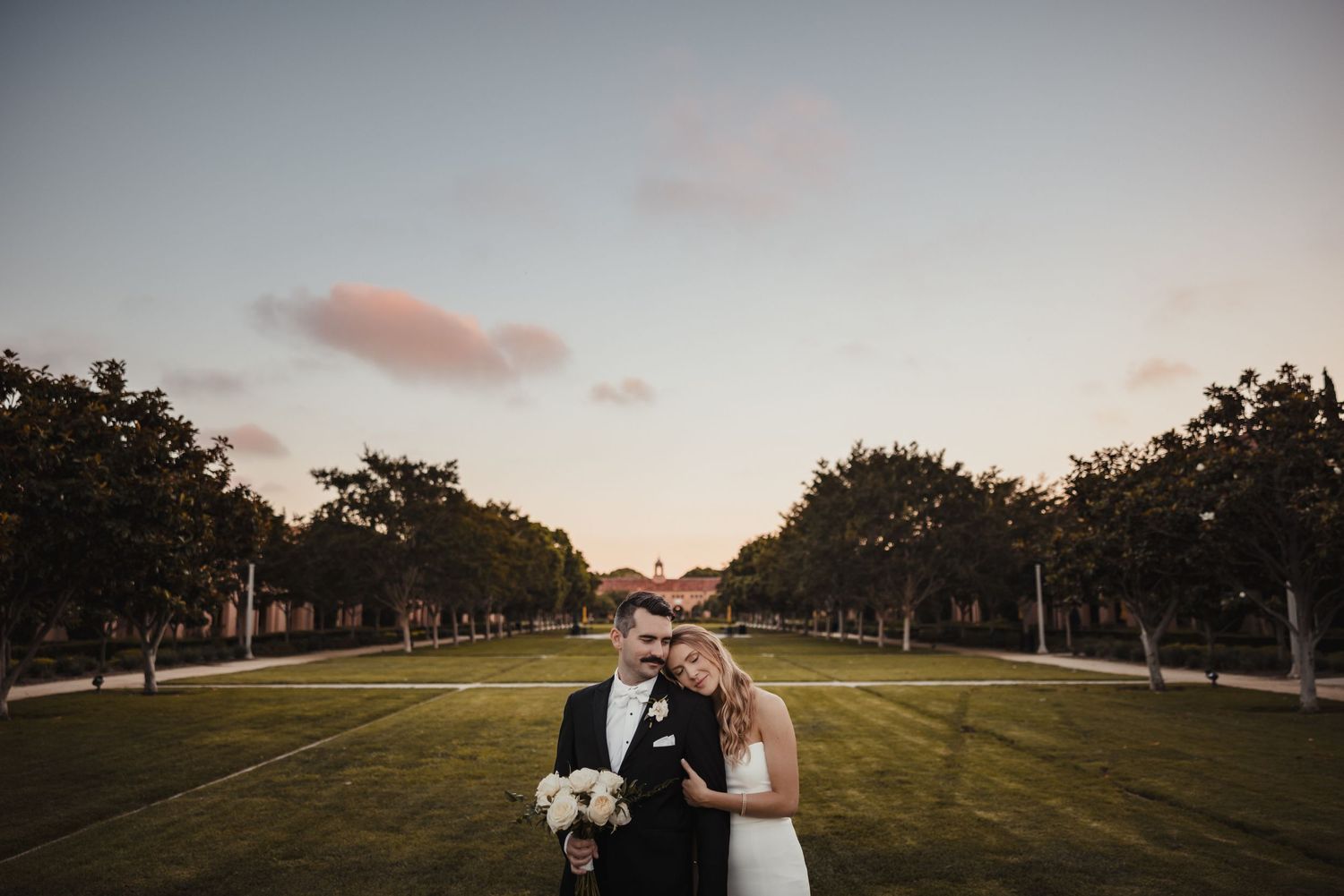 A couple stands together at sunset on a grassy lawn lined with trees holding a bouquet and beverage.
