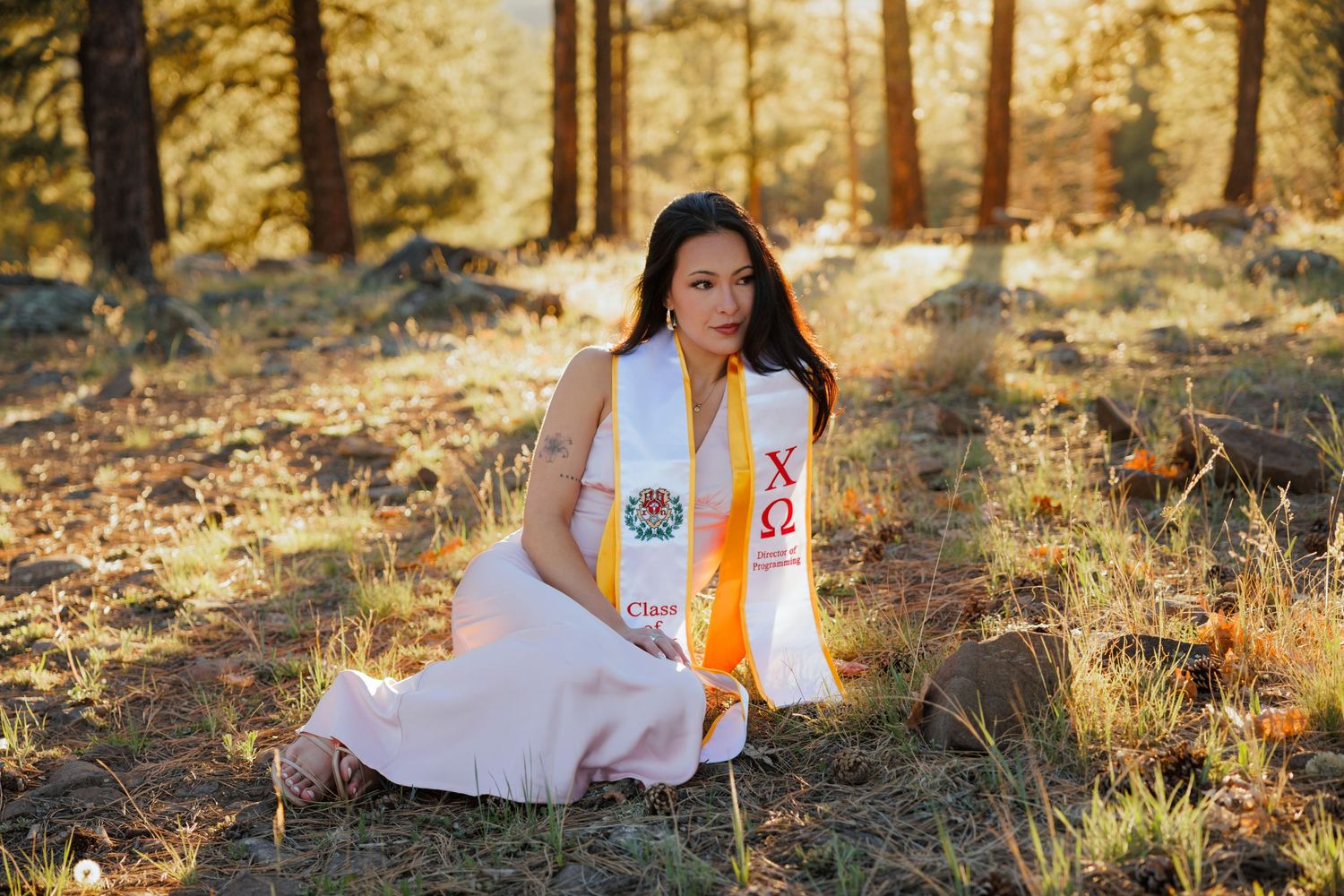 A young woman in a white Chi Omega graduation stole sits gracefully in a sunlit forest clearing.
