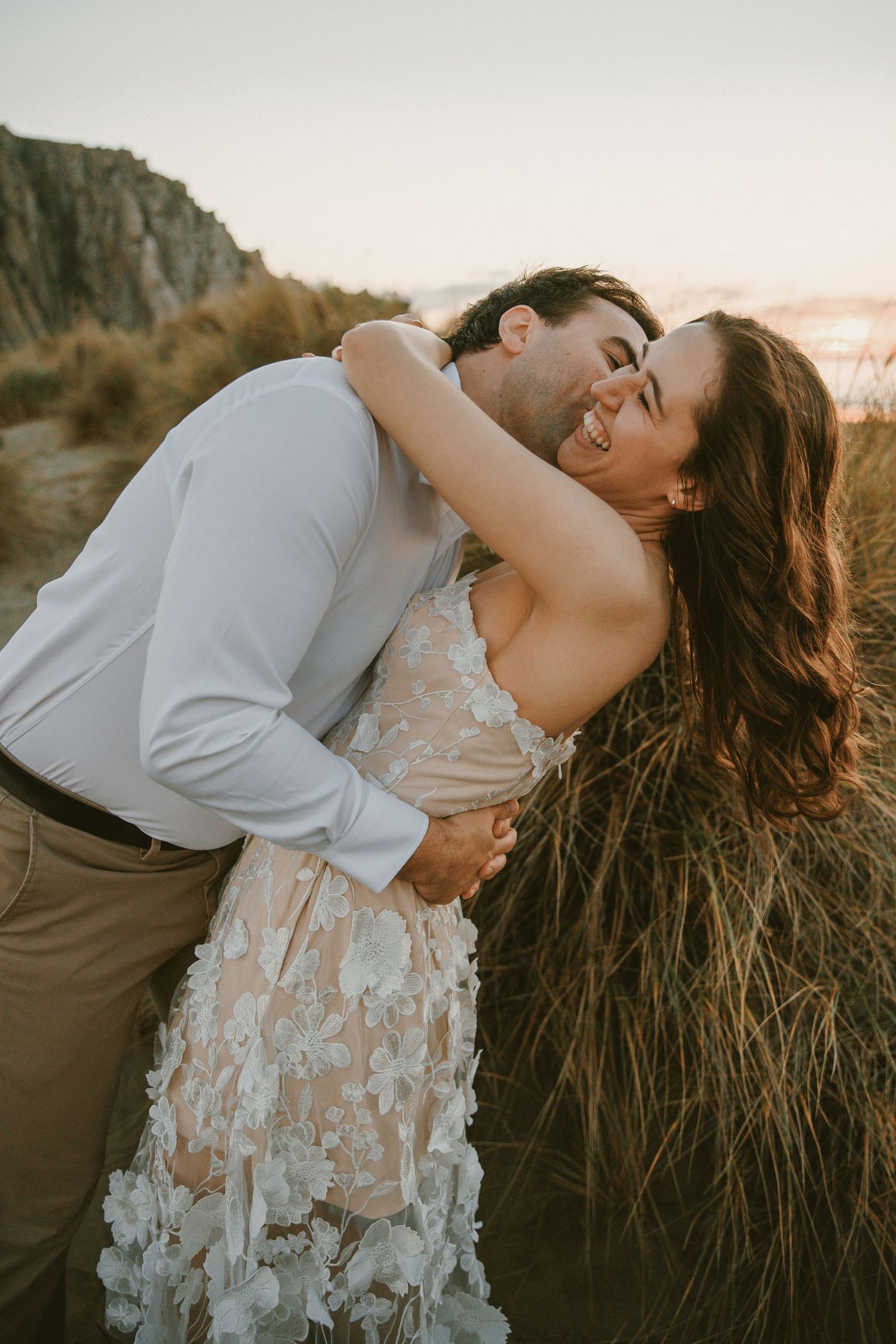 Groom kissing bride on the neck as she laughs joyfully in a floral wedding dress.