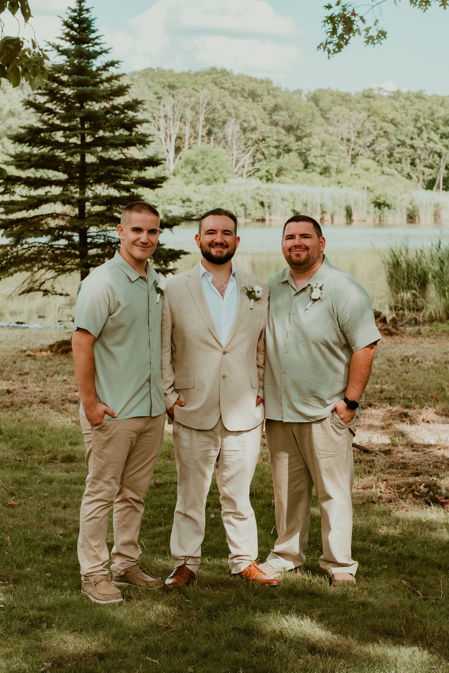 Three men in casual attire pose together by a lake with pine trees in the background.