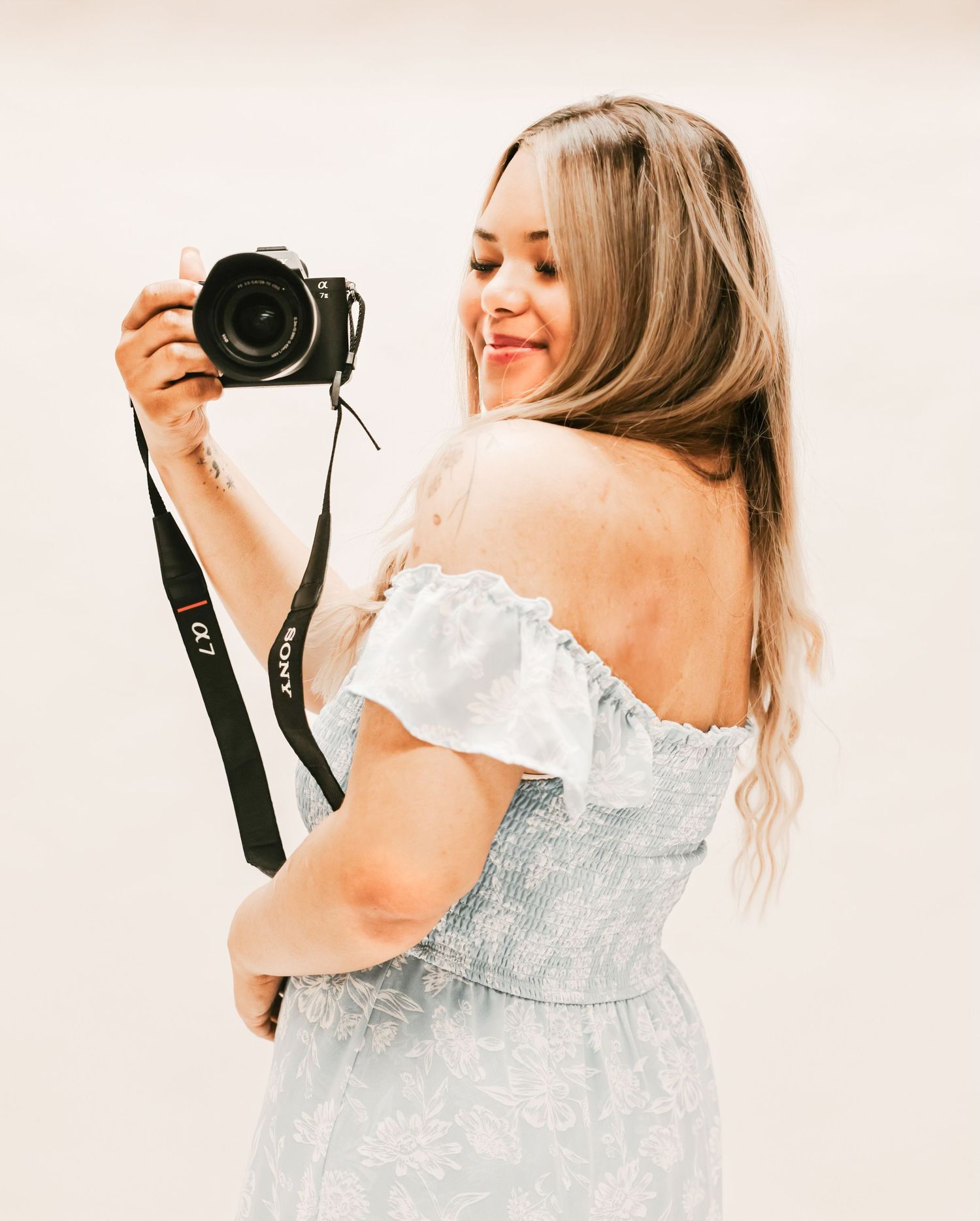 A photographer in an off-shoulder white dress holds a camera while looking over her shoulder.