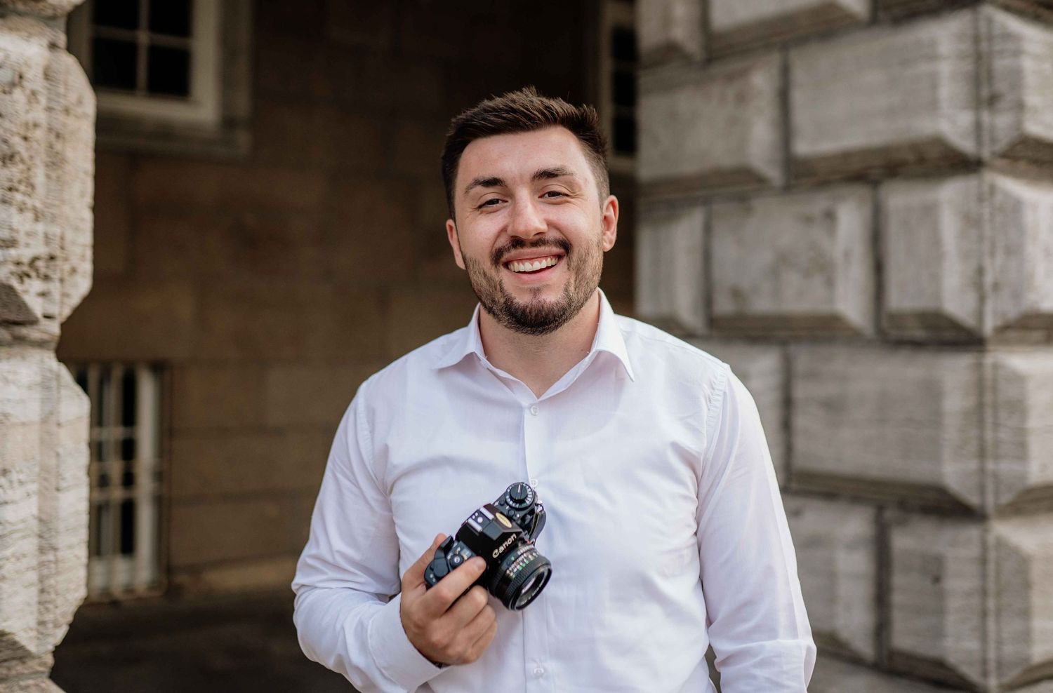 Professional photographer in a white shirt holding a camera against a stone building backdrop.